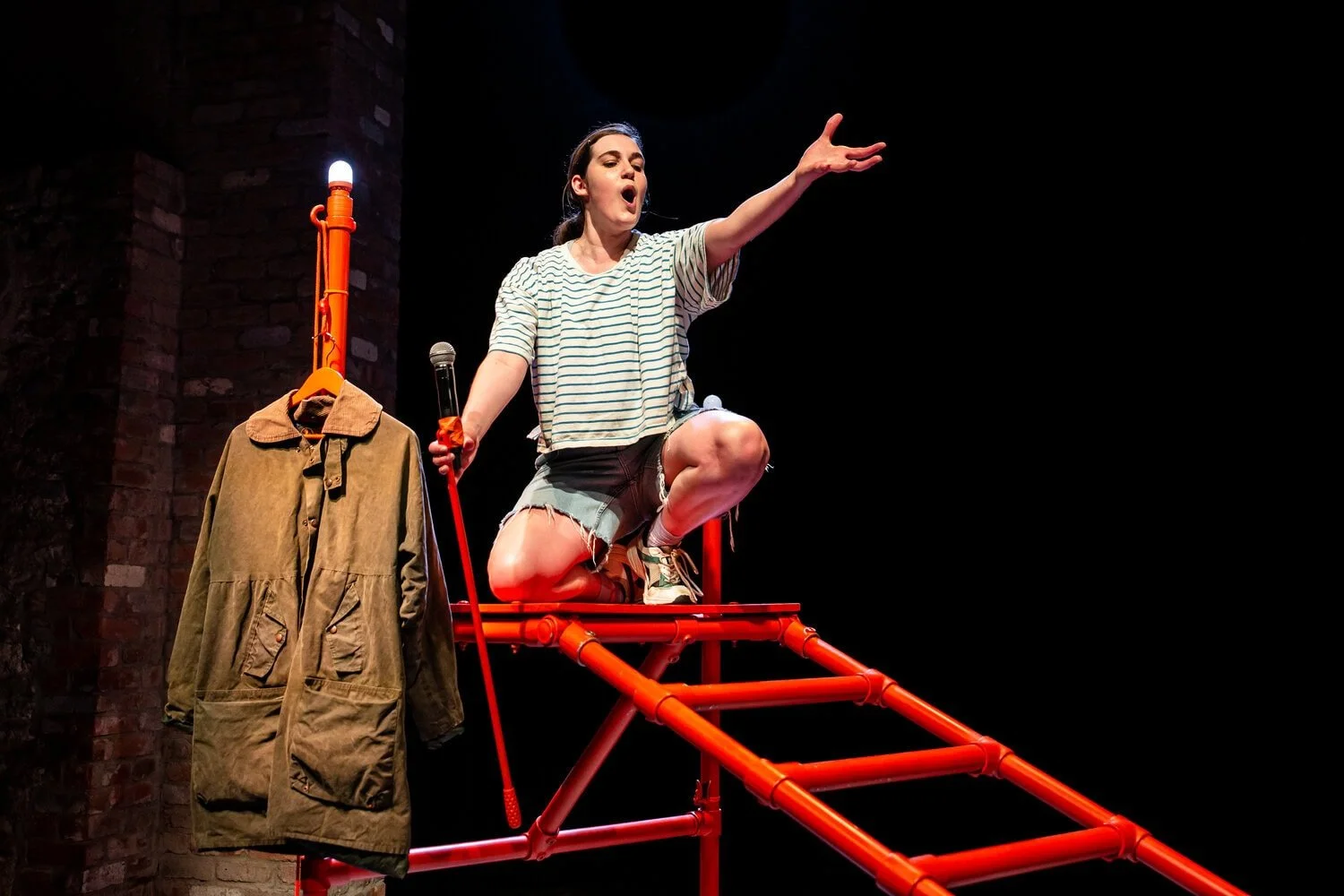 Saffi kneels on the orange frame with microphone in one hand and hand outstretched, with mouth open singing.