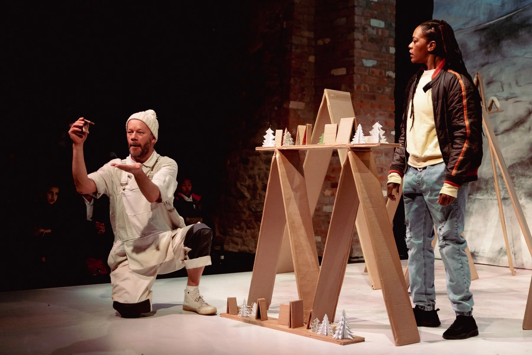 Belle kneels on the left, in the middle are cardboard minature trees and houses. Sebastien stands behind the cardboard structure looking at Belle.