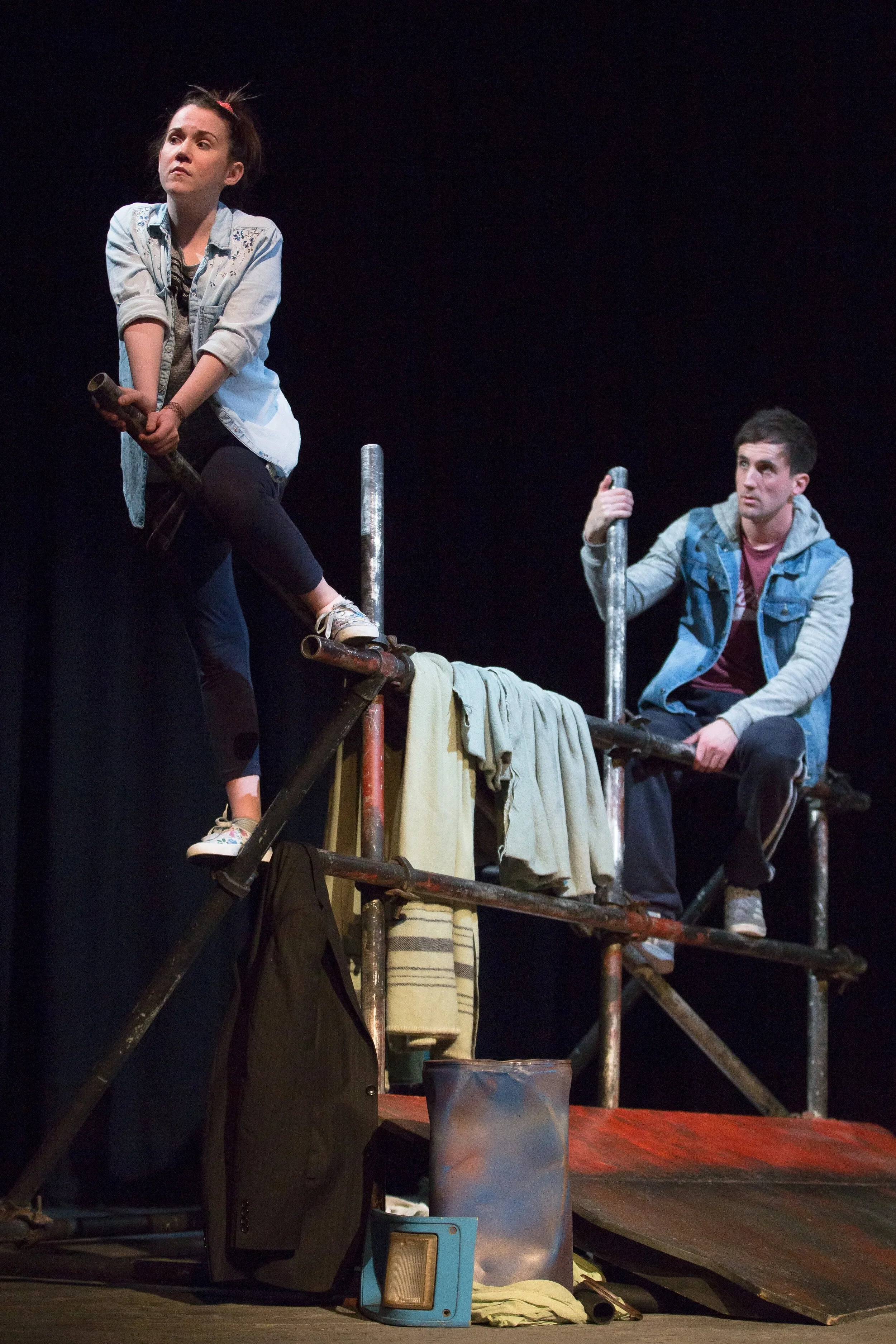 Two actors performing on stage, one seated on scaffolding and the other standing on it, with a dark background.