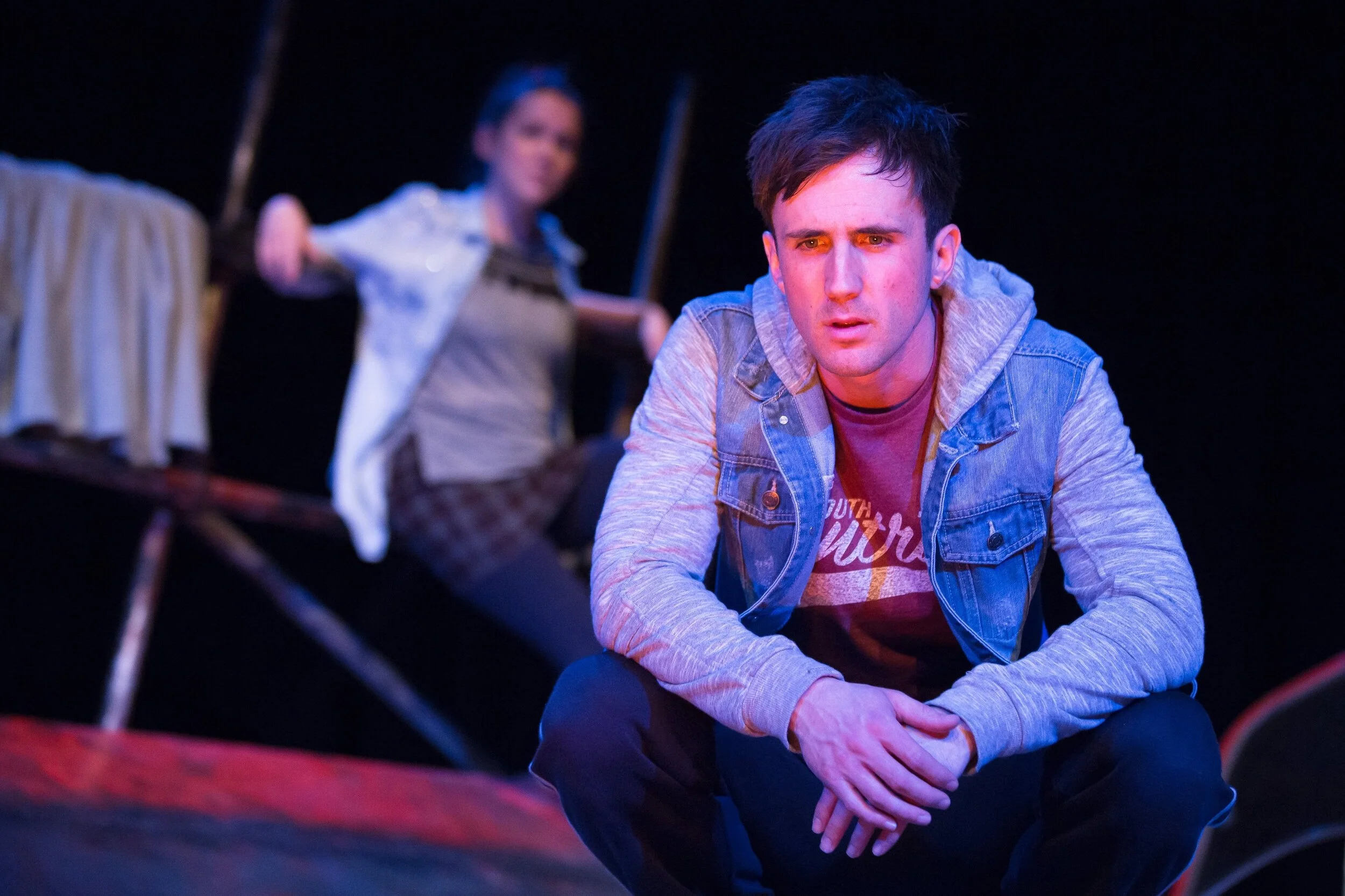 Young man crouching and looking concerned, with a woman in the background sitting on a platform and smiling, set on a dark stage.