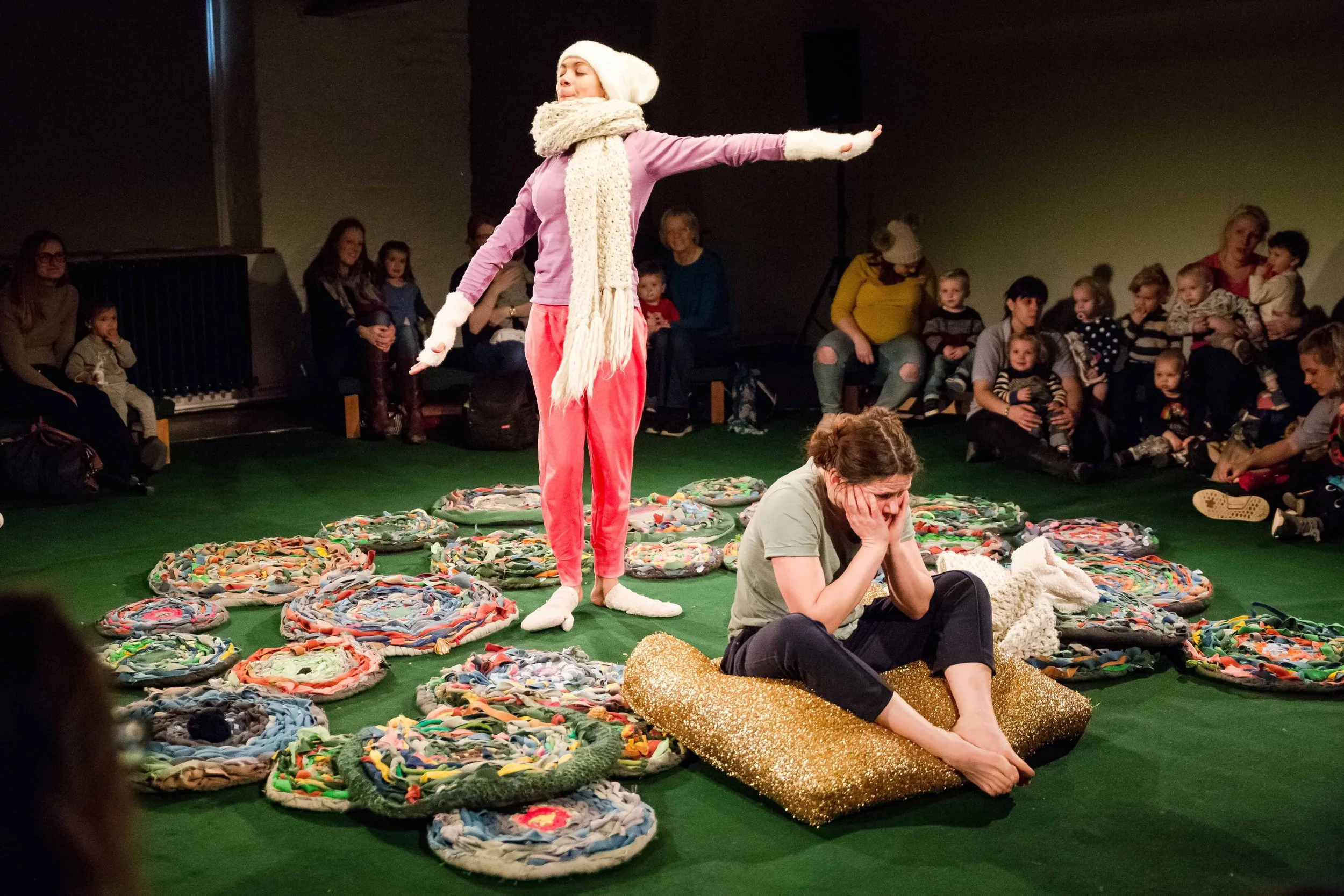 One performer sits on a cushion while the other stands behind, surrounded by cicle colourful mats. The audience sit in the background looking on.
