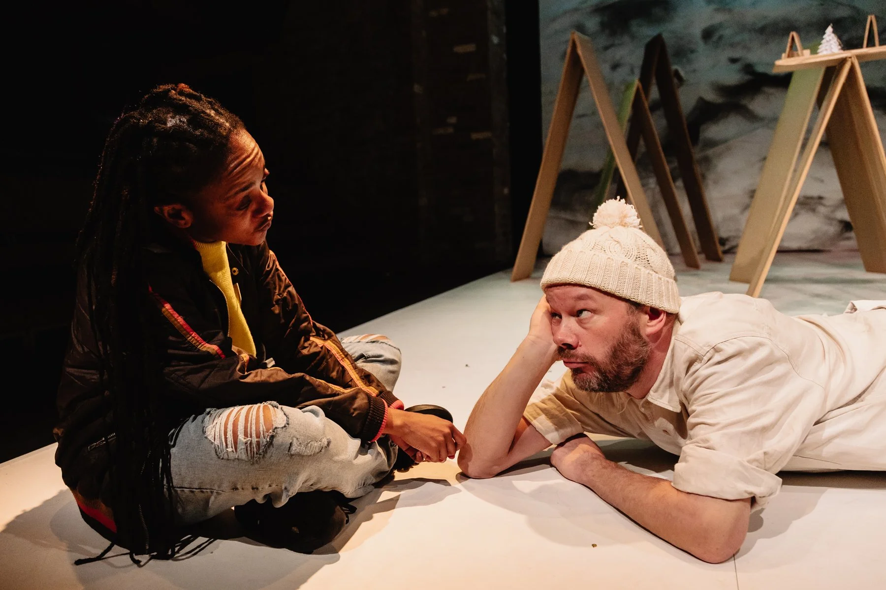 Belle lies with his head resting on his elblow looking up at Sebastien who sits cross legged on the floor looking at Belle.