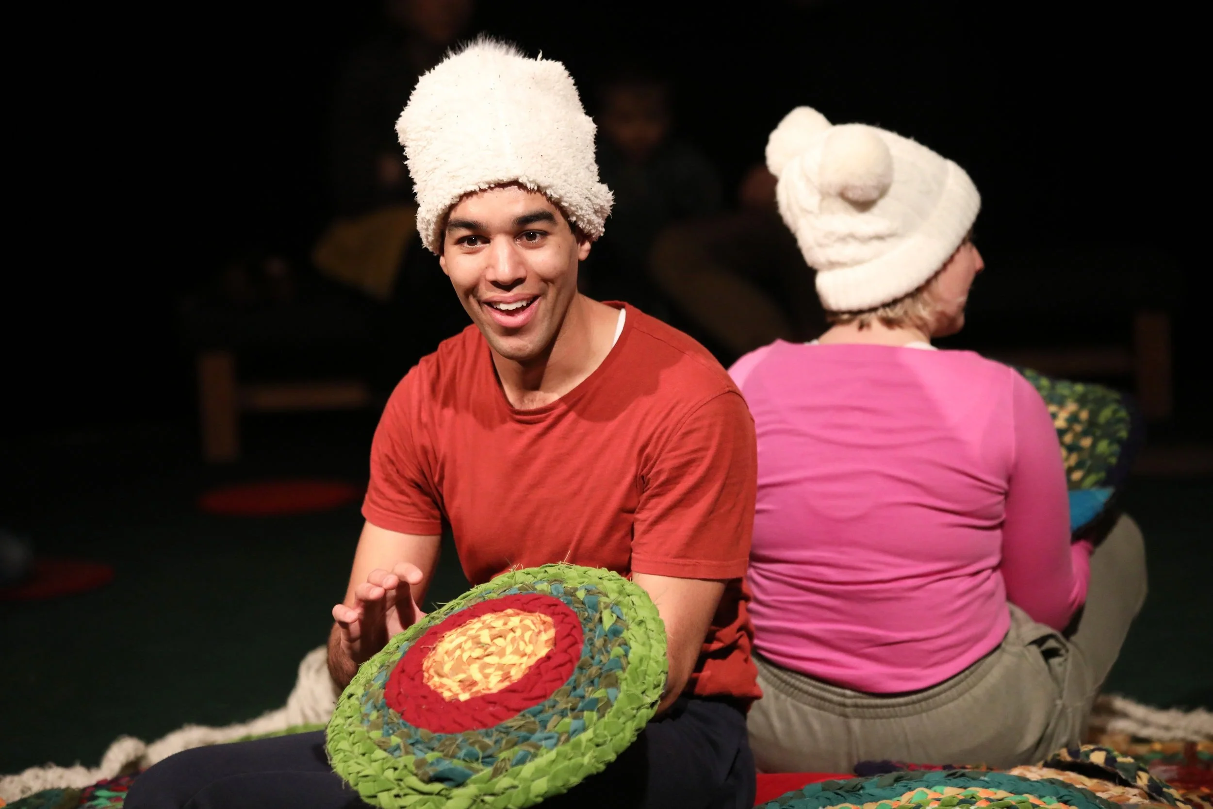 Sadie and Ira sit on a pile of colourful mats. Sadie is looking towards the camera holding a circle mat and wearing a white hat.