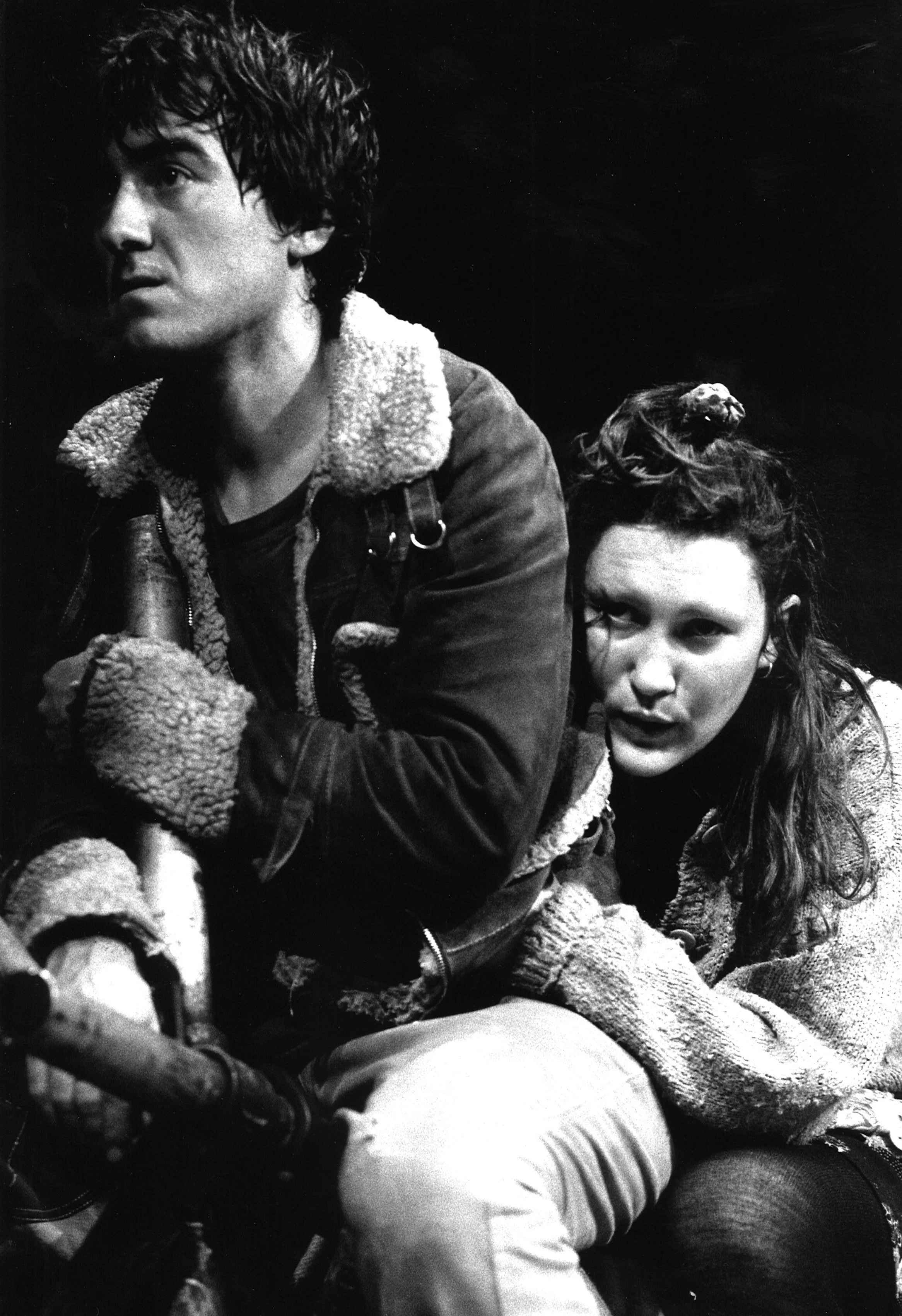 A black and white photo of a young man and woman, both with tousled hair, sitting close together. The man is holding a bicycle handlebar, and the woman is leaning on him, looking directly at the camera.
