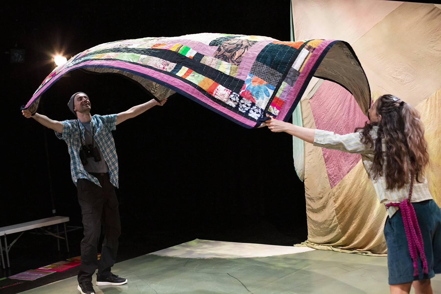 Two people are holding a colourful quilt, and they are in a dark room with a backdrop of large, textured fabric curtains in pastel colours.