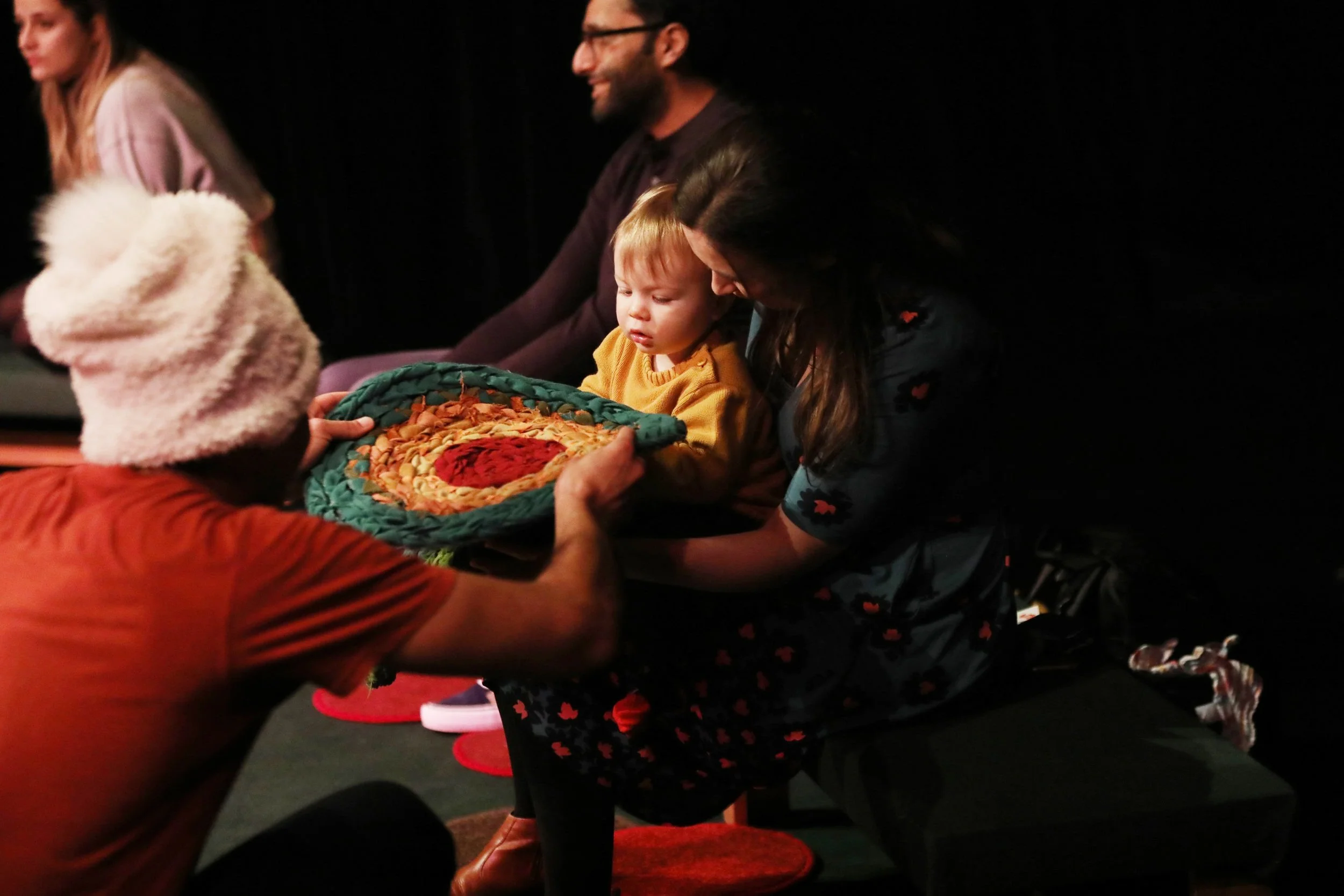 Sadie hands a colourful mat to a child in the audience who is sitting on their mother's lap.