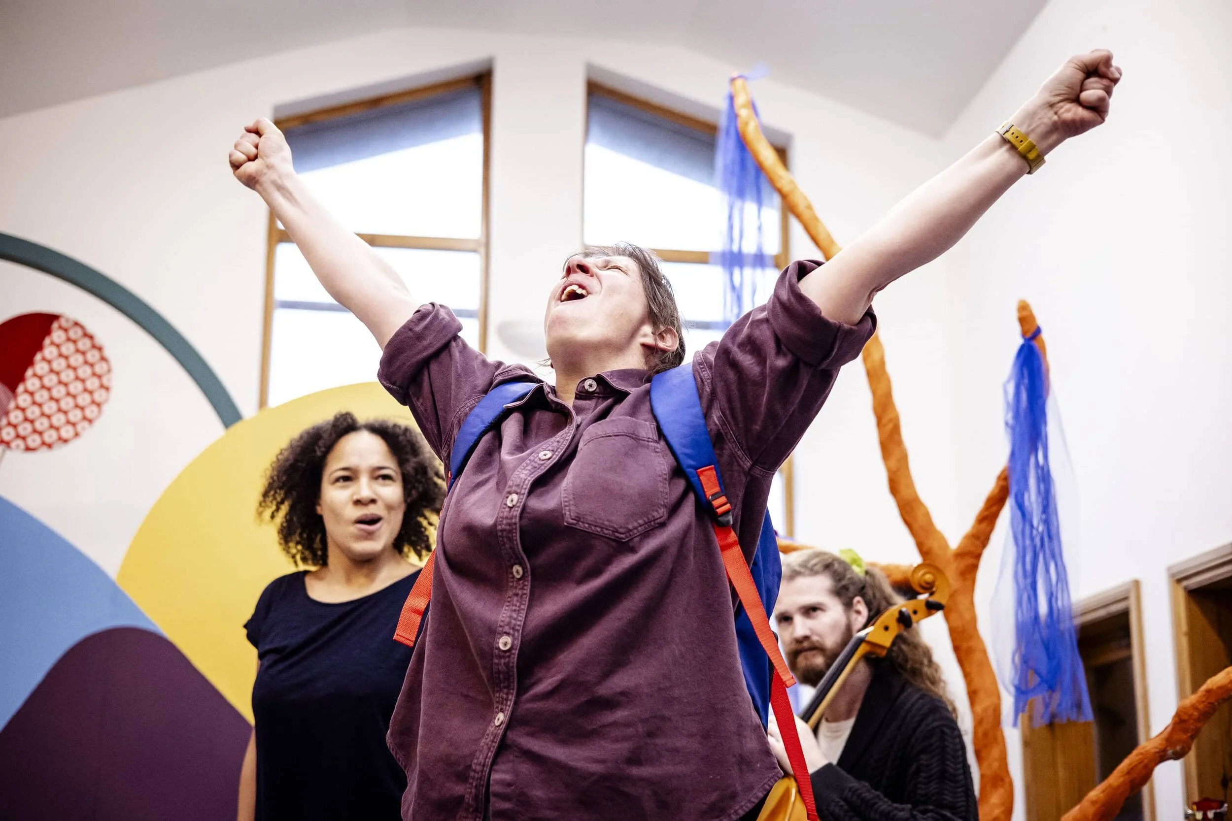 Amy stands in the foreground with her arms stretched out above her. Remi and James look on in the background. James is sitting and playing a cello.