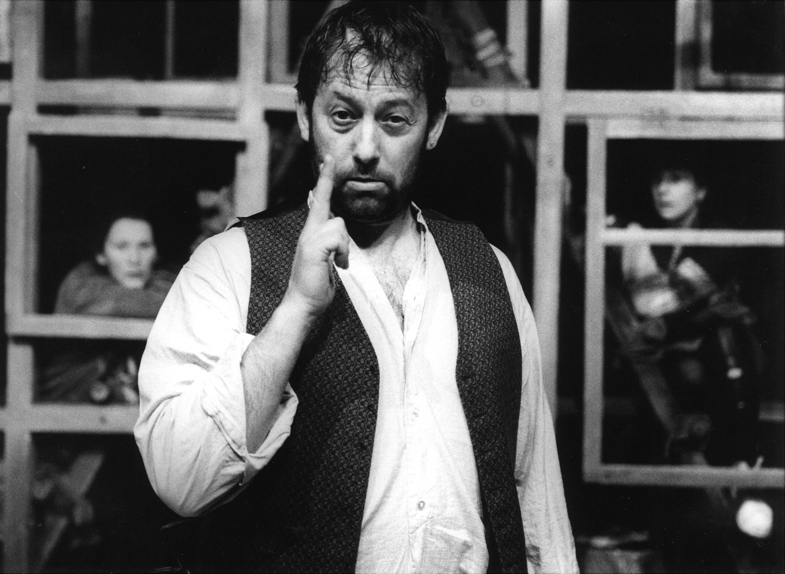 Black and white photo of a man with messy hair and a beard, wearing a vest and shirt, with his hand near his face.