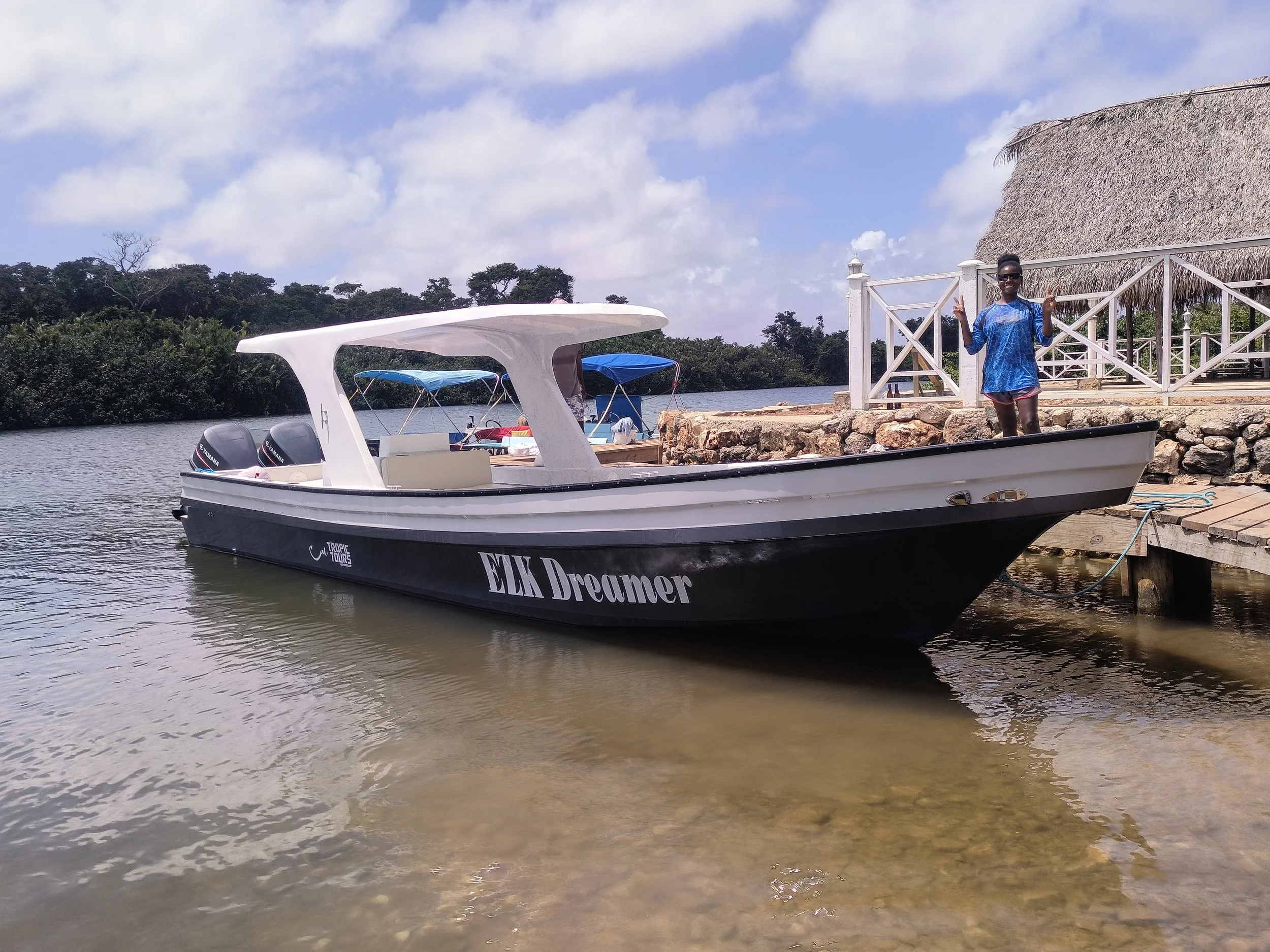 A woman standing on a dock next to a black and white boat named 'ELK Dreamer' on a river, with trees and a cloudy sky in the background.