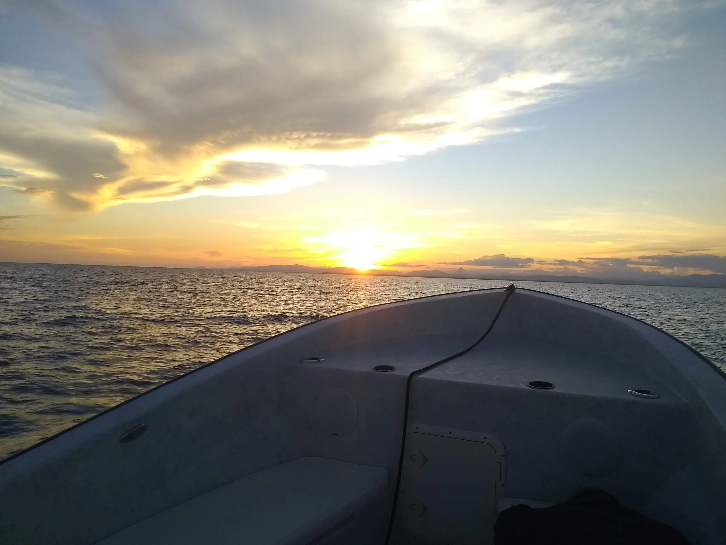 View from a boat at sunset, showing the ocean with waves and a sky with clouds illuminated by the setting sun.