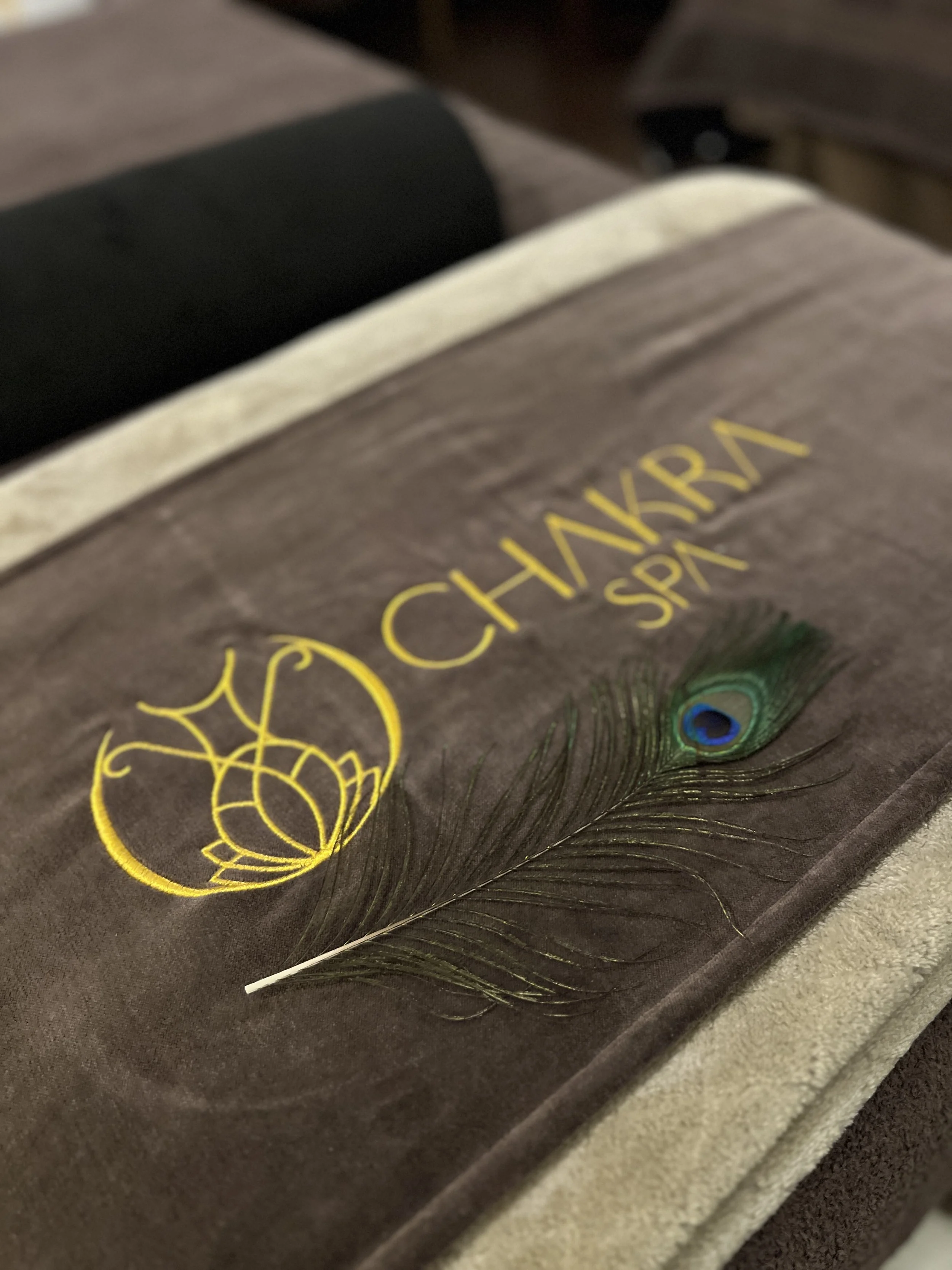 A brown towel with yellow embroidered text reading 'Chanhara Spa' and an embroidered peacock feather on it.