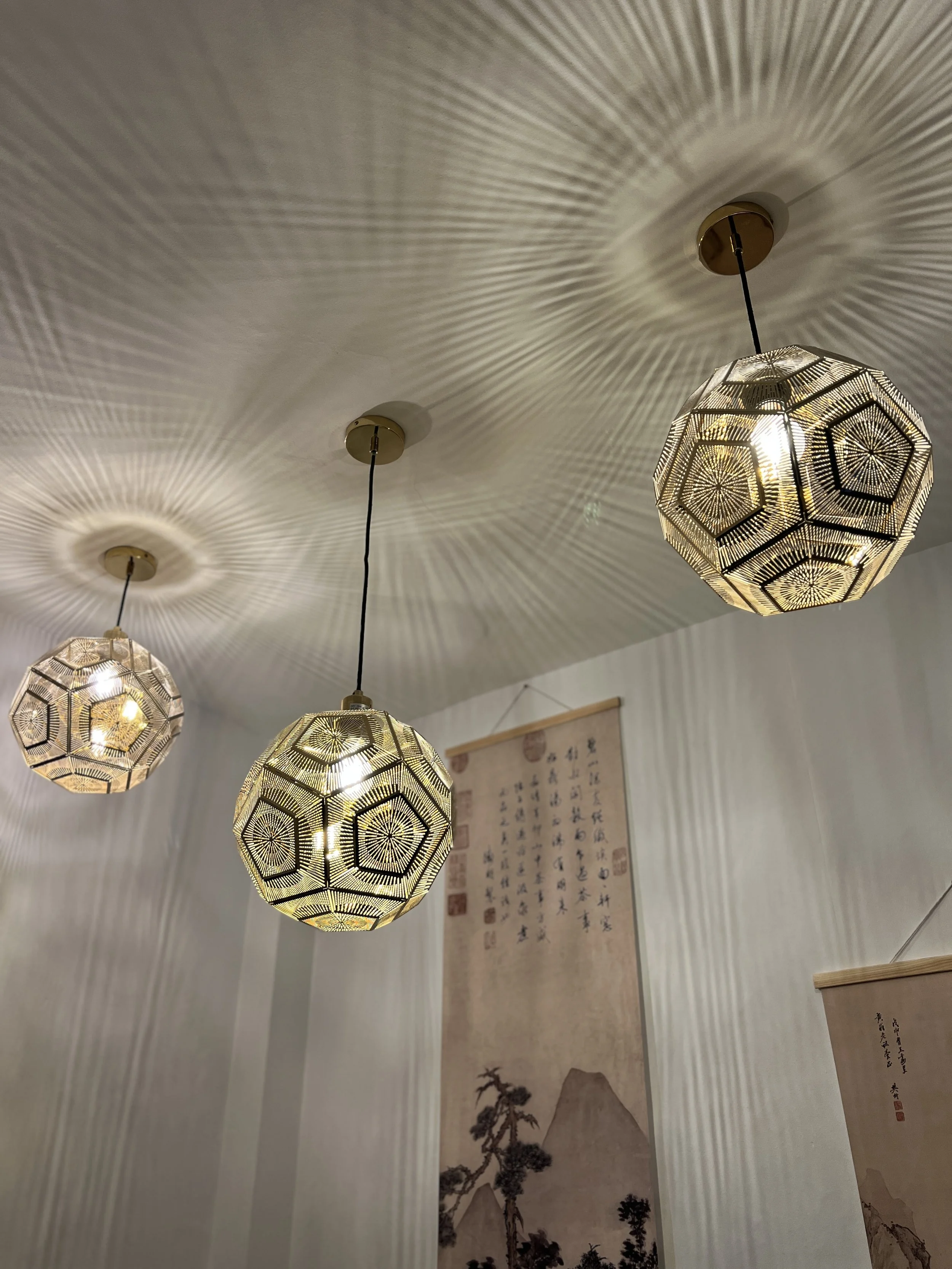 Three decorative pendant lights hanging from a ceiling, casting intricate shadows on the white ceiling. Two hanging scrolls with traditional Asian art and calligraphy are visible in the background.