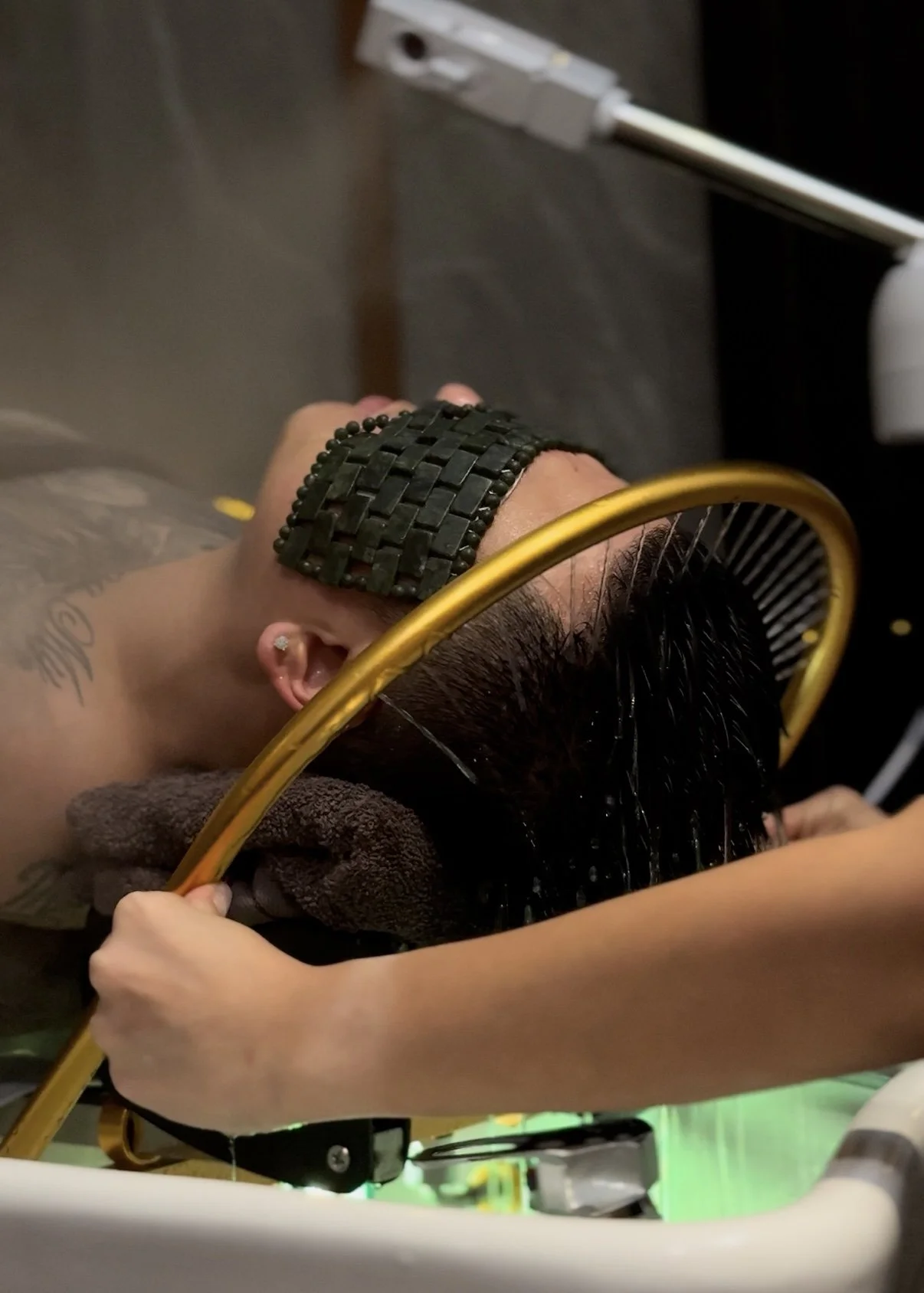 Person receiving a hair treatment at a salon, laying on a towel with hair being combed, wearing a black wristwatch, and visible tattoos on arm.