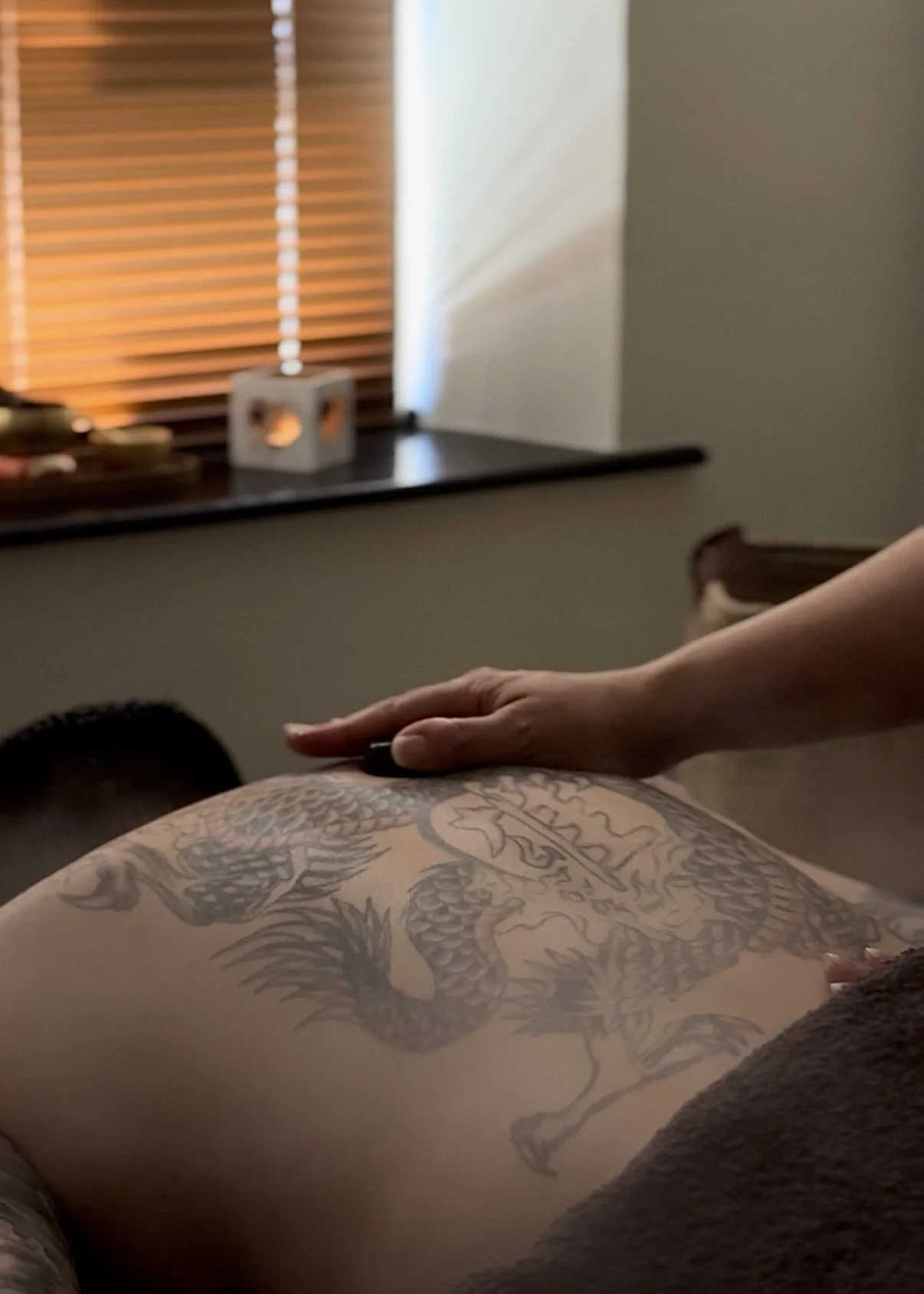 Close-up of a tattooed person's back with a dragon design while a hand rests on their back in a room with wooden blinds and soft lighting.