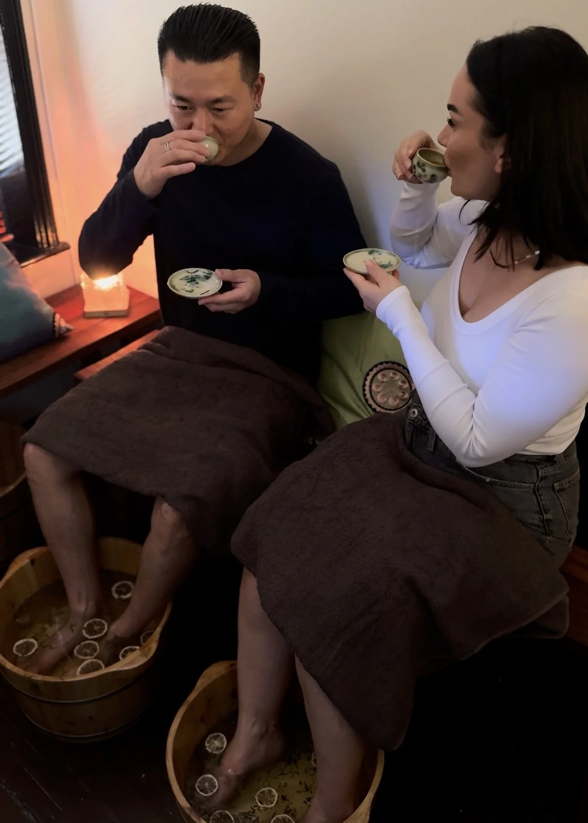 Two people sitting on a bench, soaking their feet in foot baths, while wearing brown towels around their waists, and drinking tea from ceramic cups.