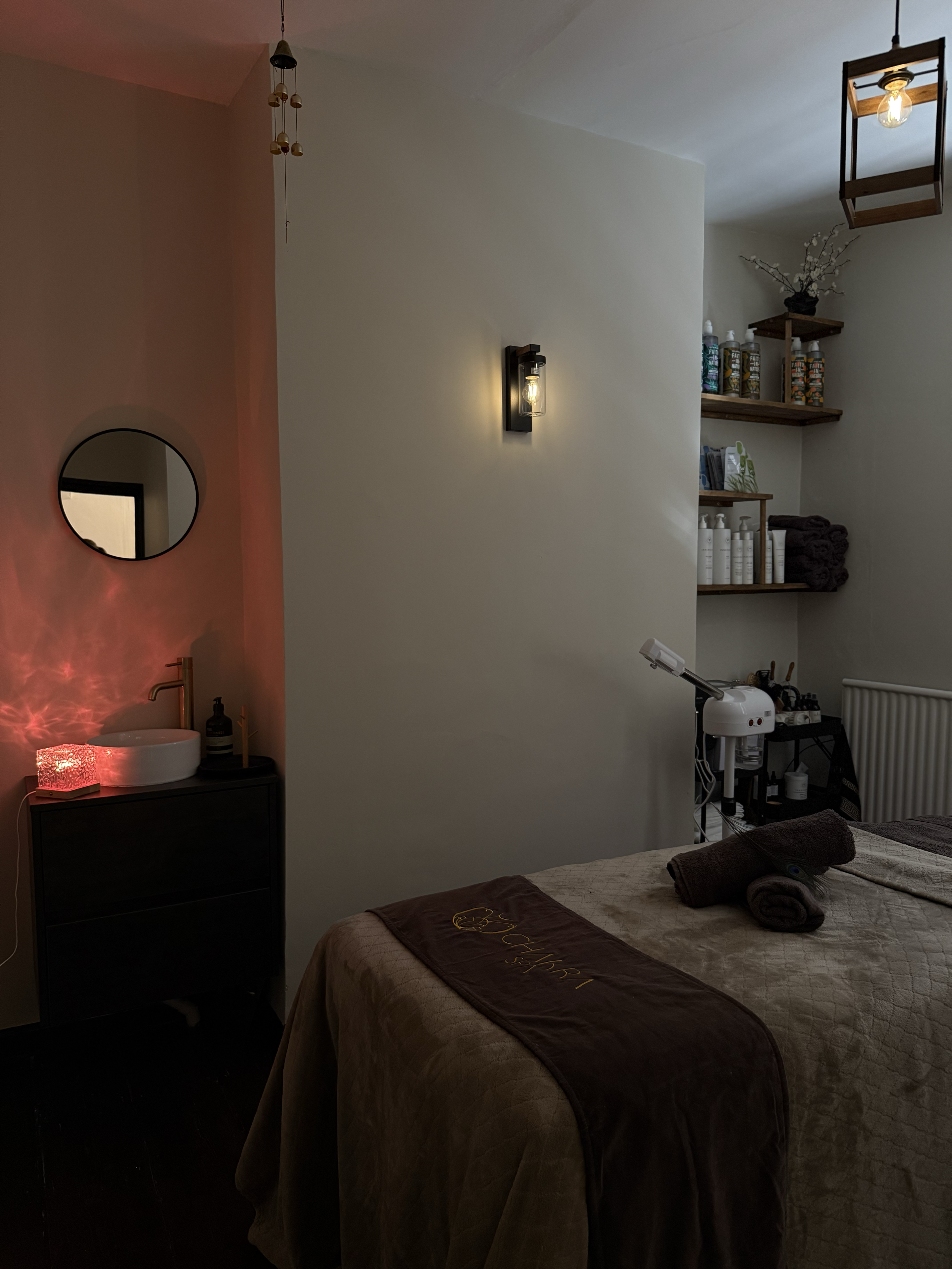A cozy massage room with a massage bed covered in a beige blanket with a brown towel on top that has the words 'Luxury Spa' embroidered on it. There are rolled towels on the bed. To the right, a massage equipment machine is visible, along with wooden