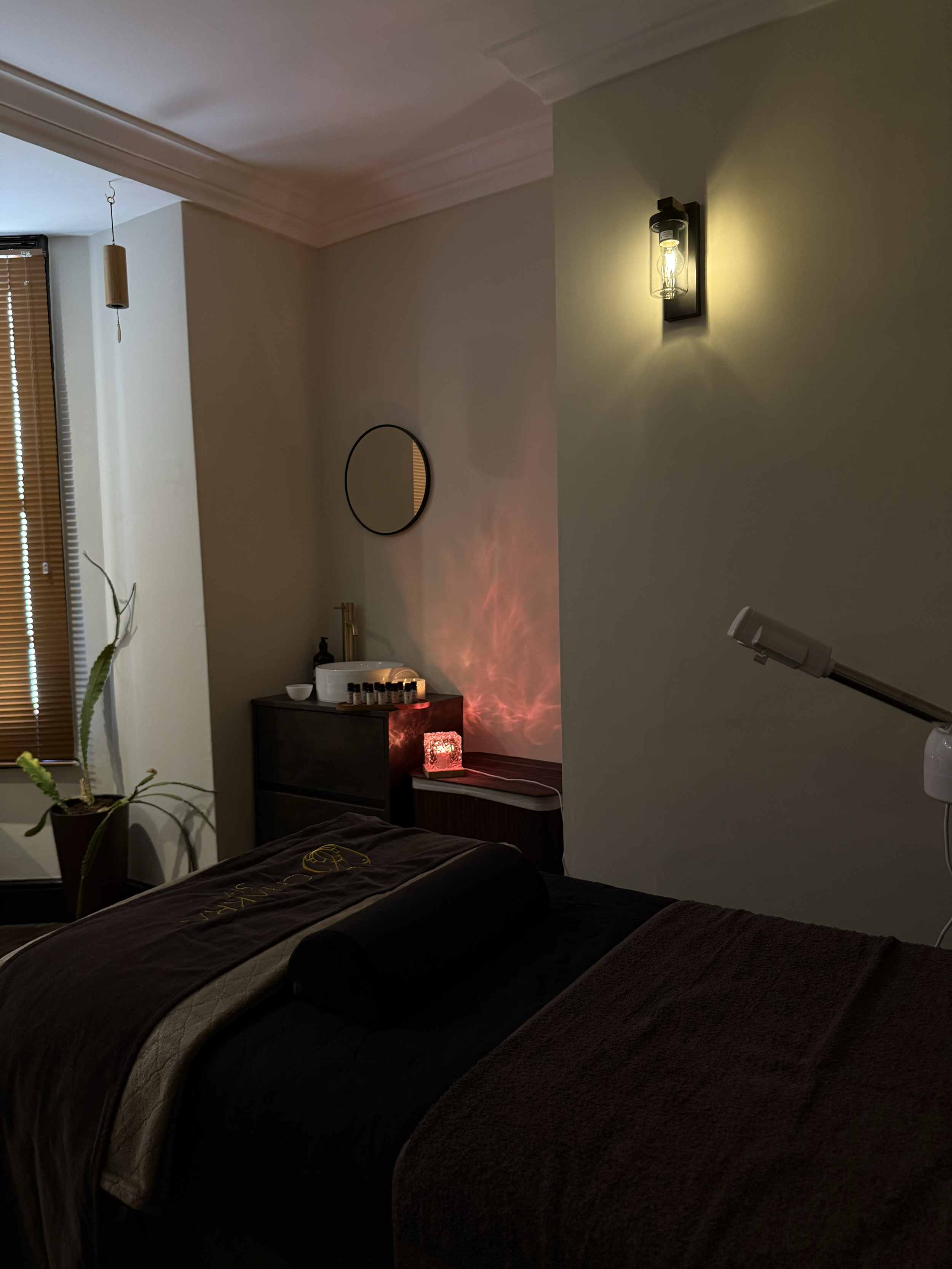 A cozy massage room with a massage table covered in dark and beige blankets, a small side table with a Himalayan salt lamp, essential oils, and a sink. The room has warm lighting, a potted plant, and window blinds.