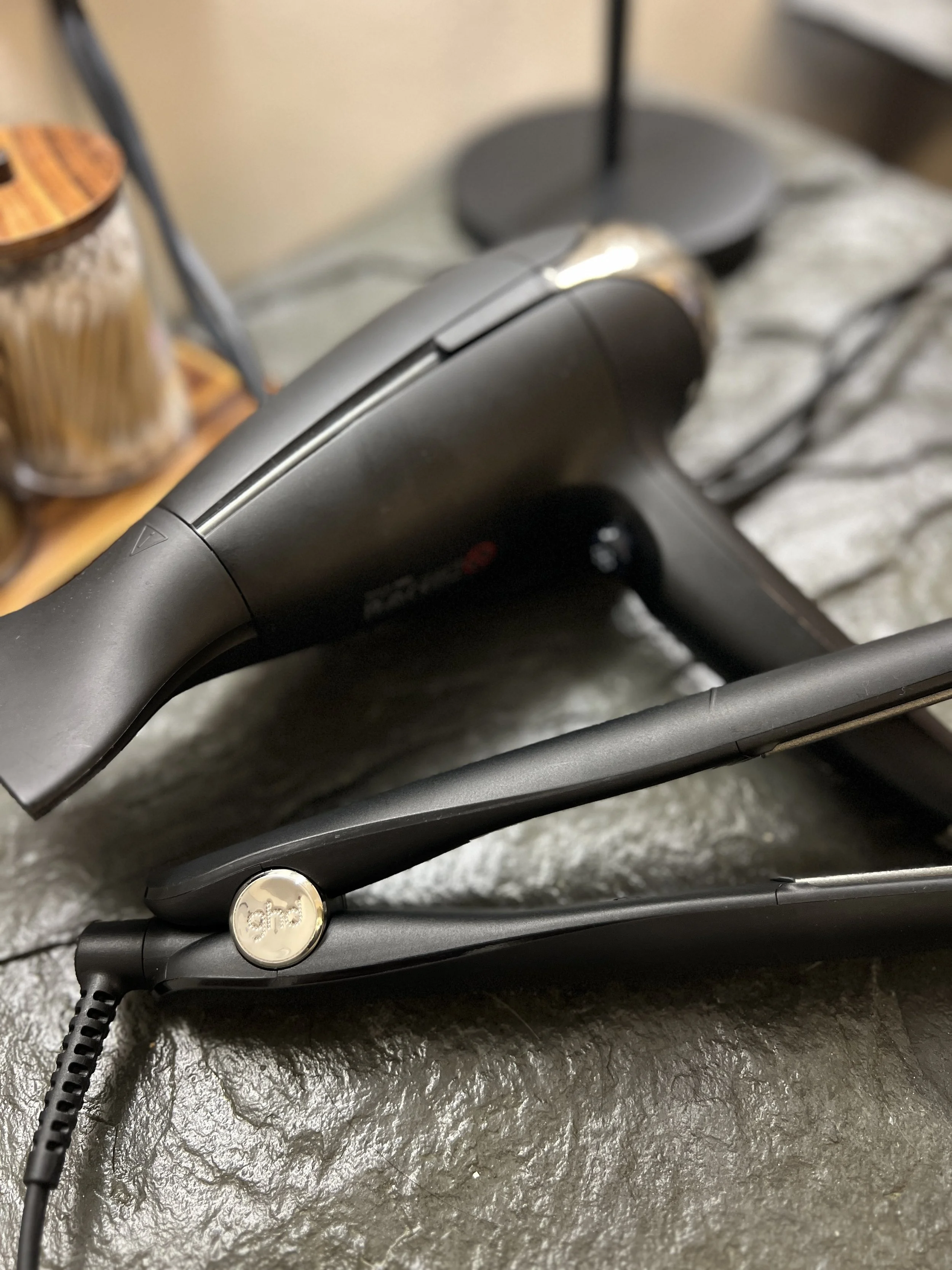 A flat iron hair straightener lying on a textured surface with a lamp and jars in the background.