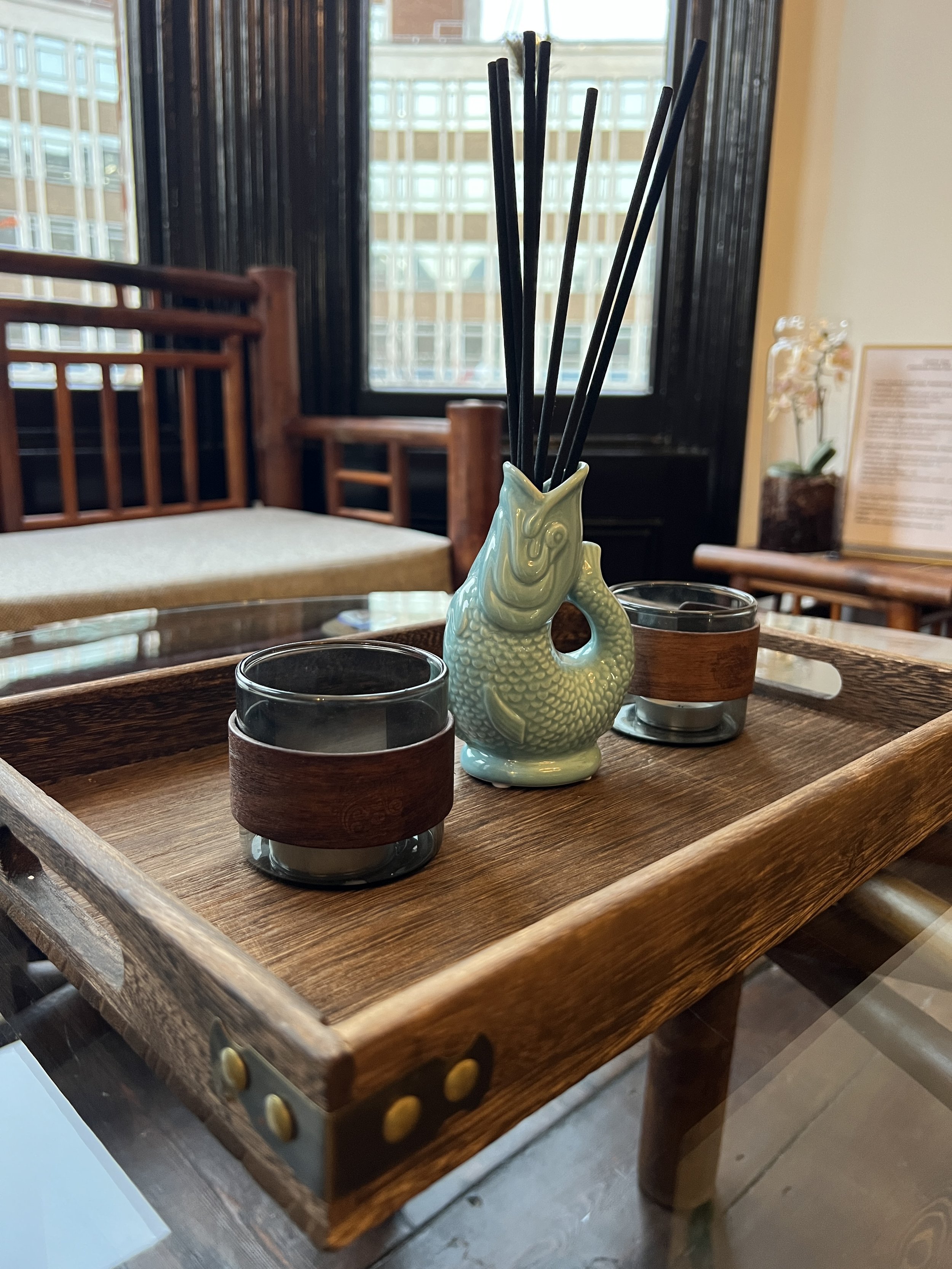 A wooden serving tray with two glass cups wrapped in wooden holders and a ceramic fish-shaped container holding black chopsticks, placed on a glass table in a restaurant with wooden furniture and large windows.