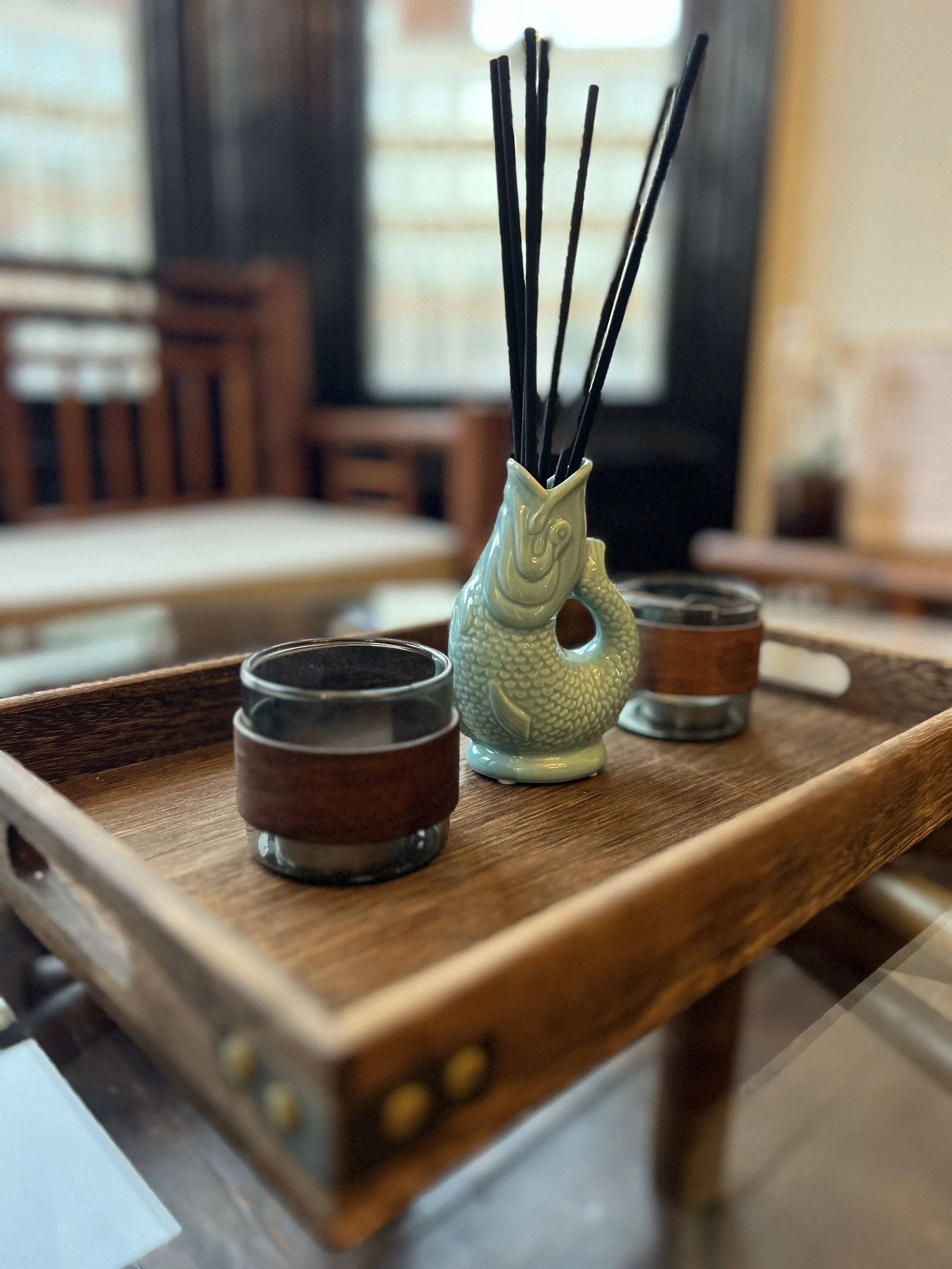 A small wooden table with a decorative porcelain fish-shaped container holding incense sticks, flanked by two small glass candle holders in a cozy indoor setting.