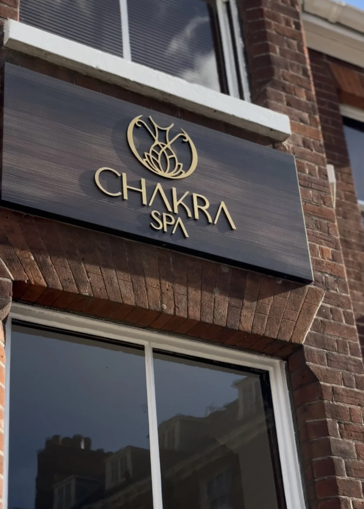 Sign for Chakra Spa attached to a brick building with a large window reflecting buildings and sky.