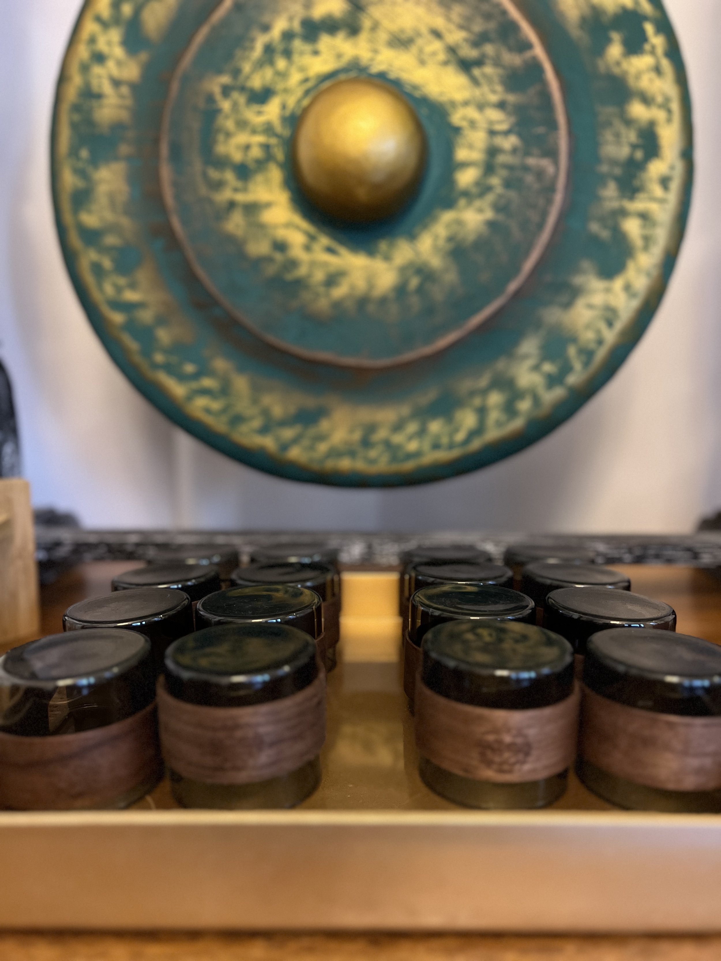 Close-up of a collection of small jars or containers with black lids arranged on a wooden surface, with a large, abstract, circular mural with yellow and green colors hanging on the wall in the background.