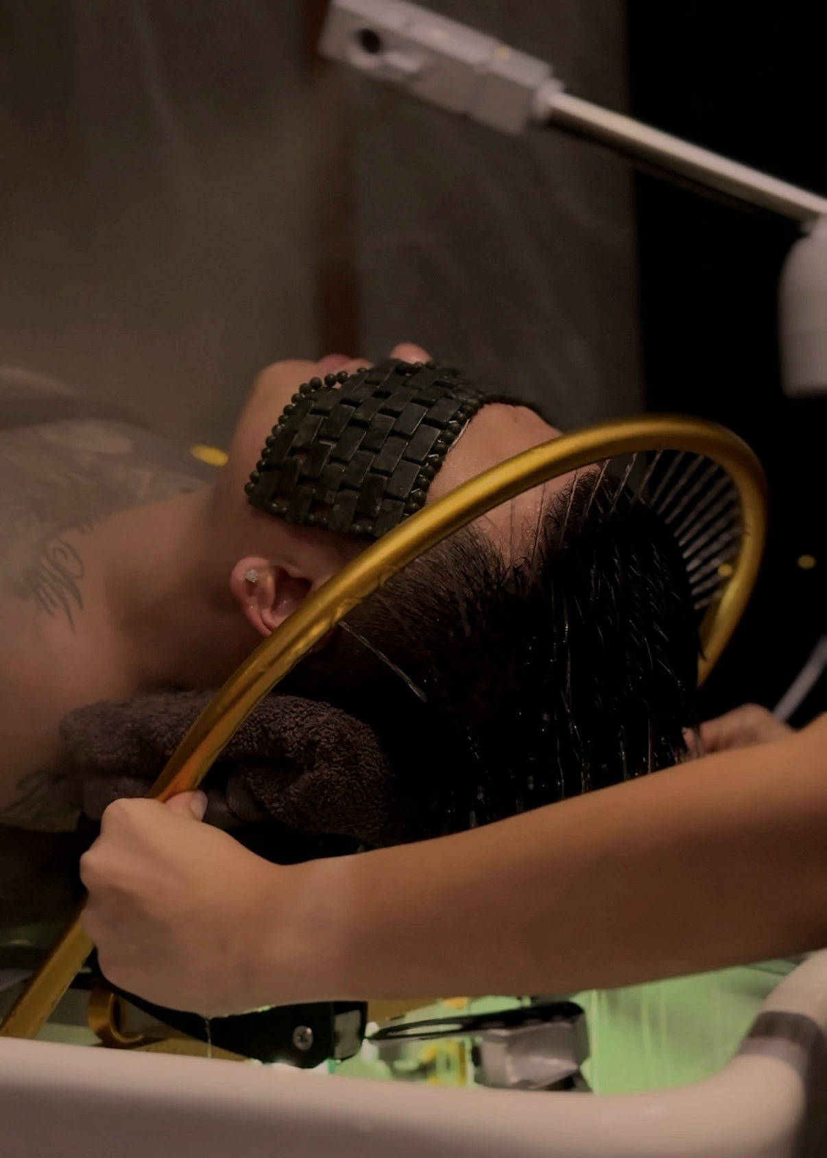 Person receiving a Japanese Head Massage, lying back with head tilted, covered with a towel, with a black woven headband and earrings visible, with a yellow hose and water streaming onto their hair.