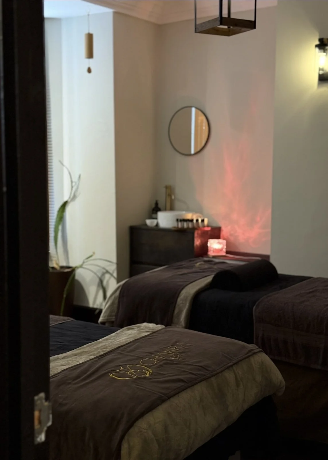 A massage or therapy room with two beds, dark brown bedding, a potted plant on the left, a small dresser with essential oils and a basin, a mirror on the wall, a salt lamp, and a red light projector casting patterns on the wall.