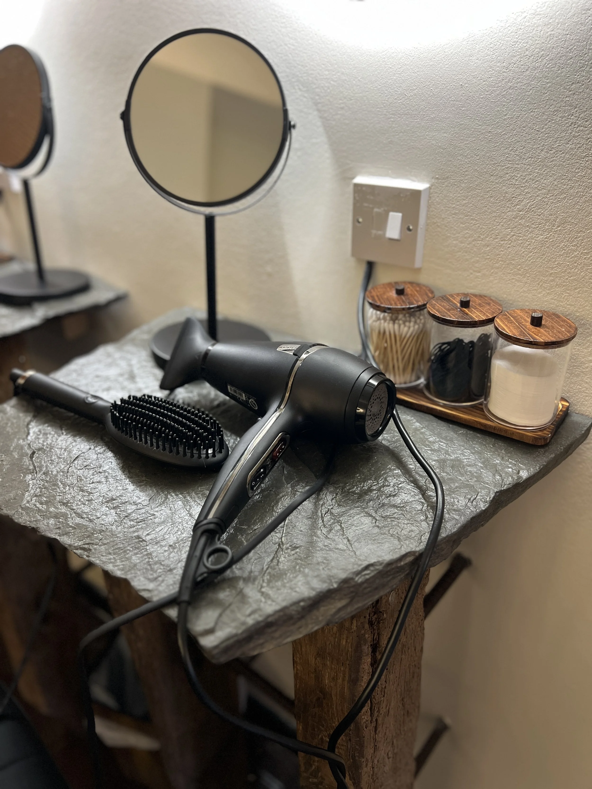 A black hair dryer and a black hairbrush on a textured gray stone tabletop, with small jars of hair accessories and a mirror in the background.