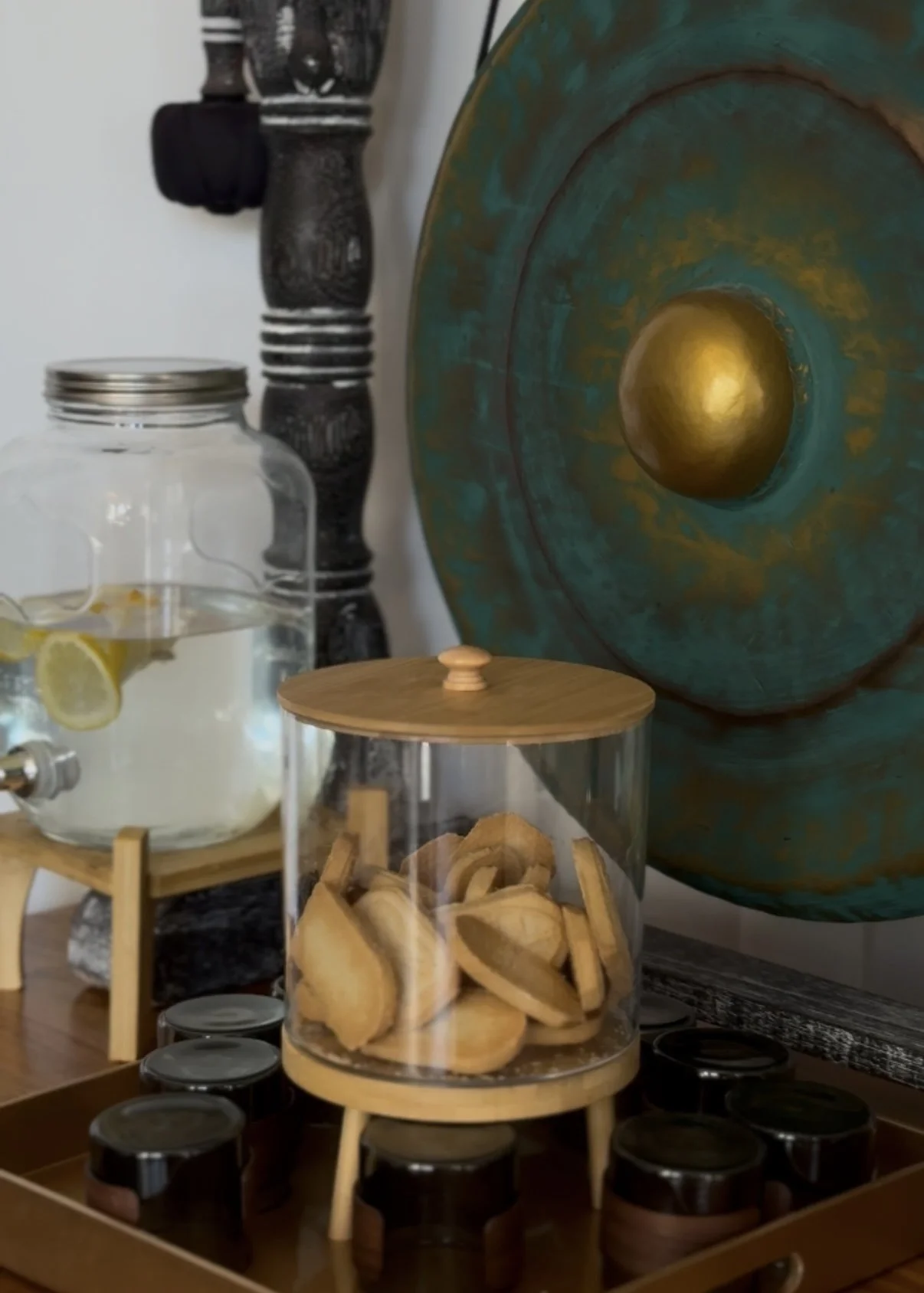 A glass jar with a lid containing lemon slices, a clear container with wooden lemon cookies, and a large round decorative wall hanging with a gold center in a room with a dark wooden table.