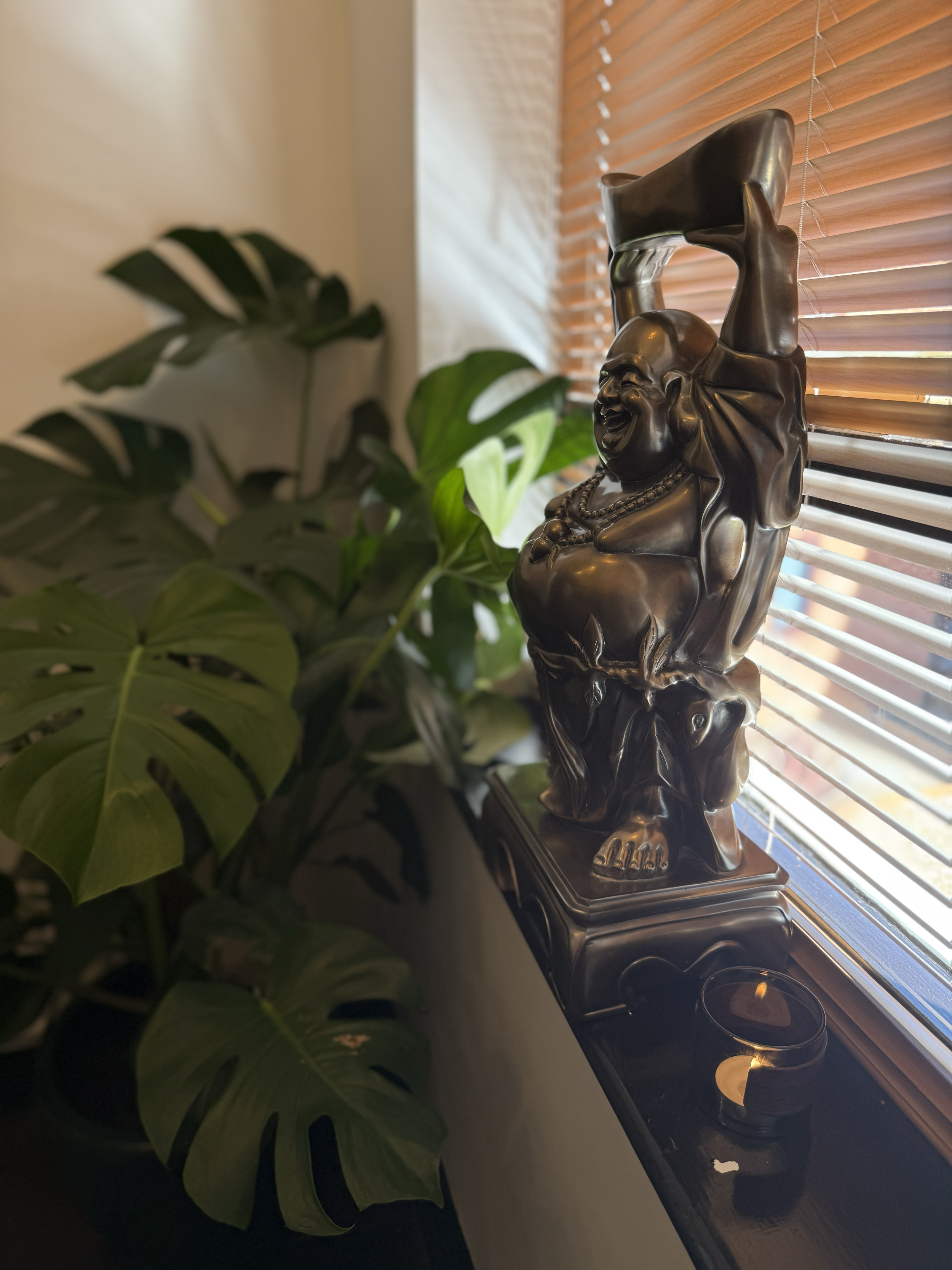 Bronze Buddha statue on a windowsill next to a Monstera plant and a lit candle in a black holder.