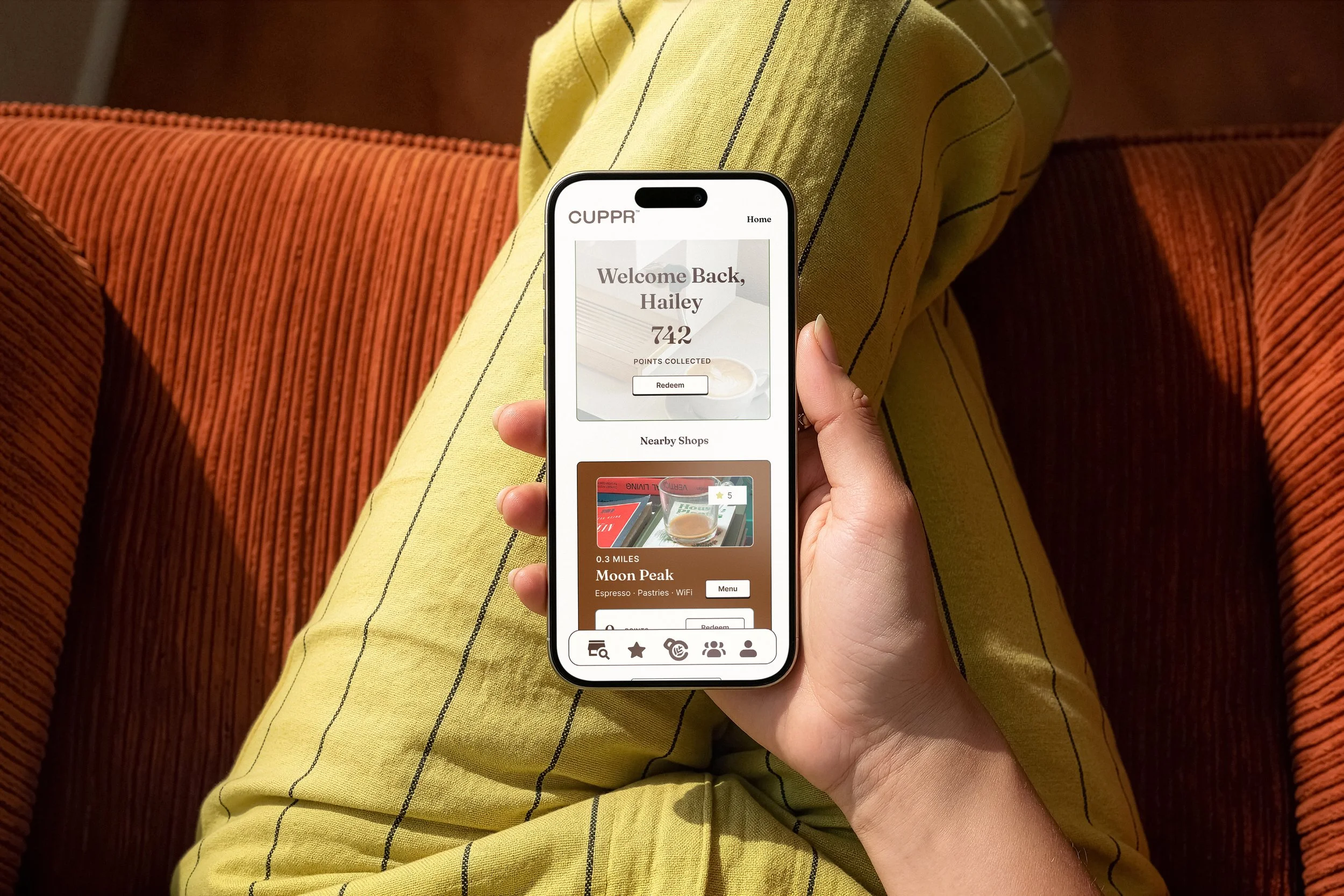 A person holding a smartphone with a rewards app displaying a welcome message and points collected, sitting on an orange striped couch with yellow and black striped pants.
