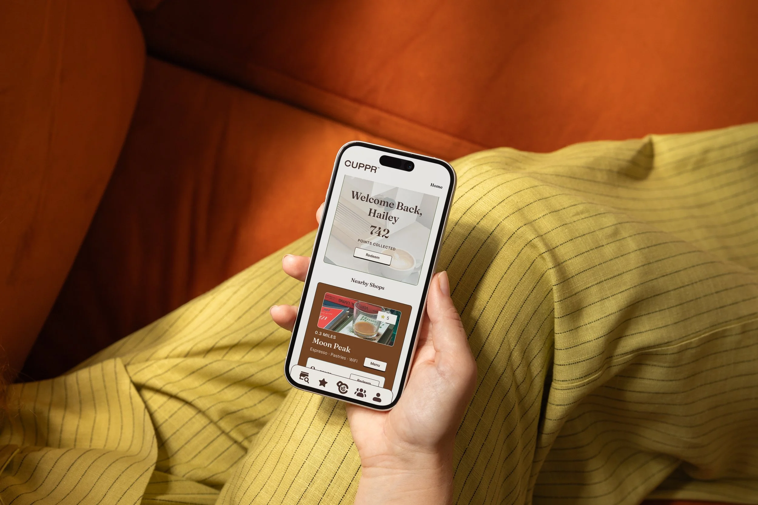 Person wearing yellow striped pants sitting on an orange couch, holding a smartphone with a loyalty app showing welcome message and points, with a yellow pillow nearby.