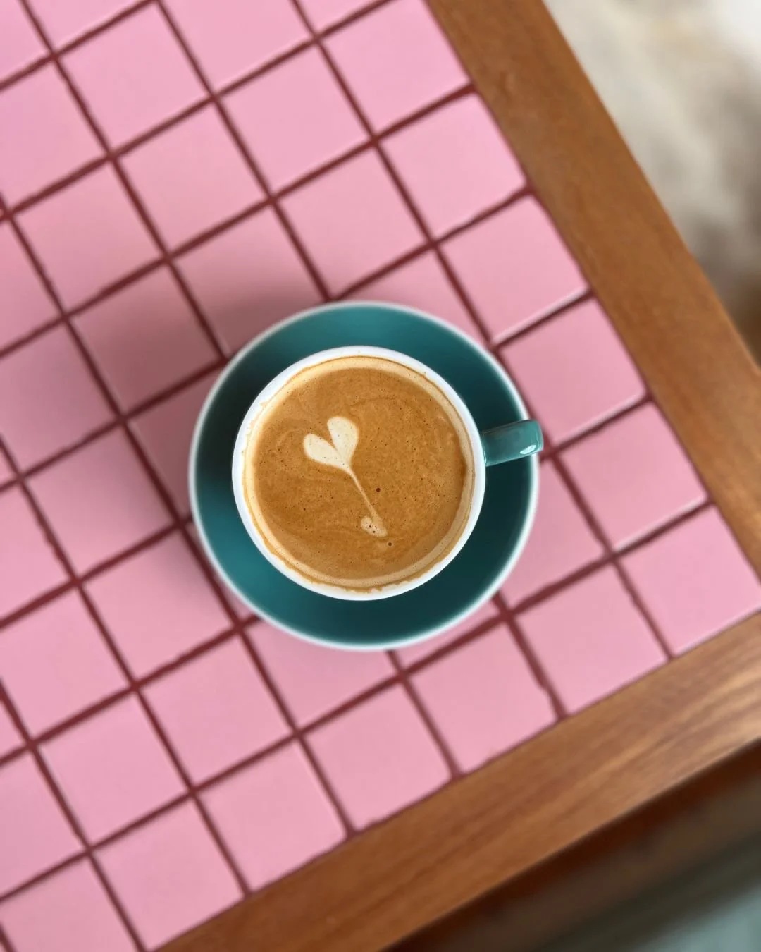 A cup of coffee with a latte art heart-shaped foam design, sitting on a teal saucer and placed on a pink tiled table with wooden edges.