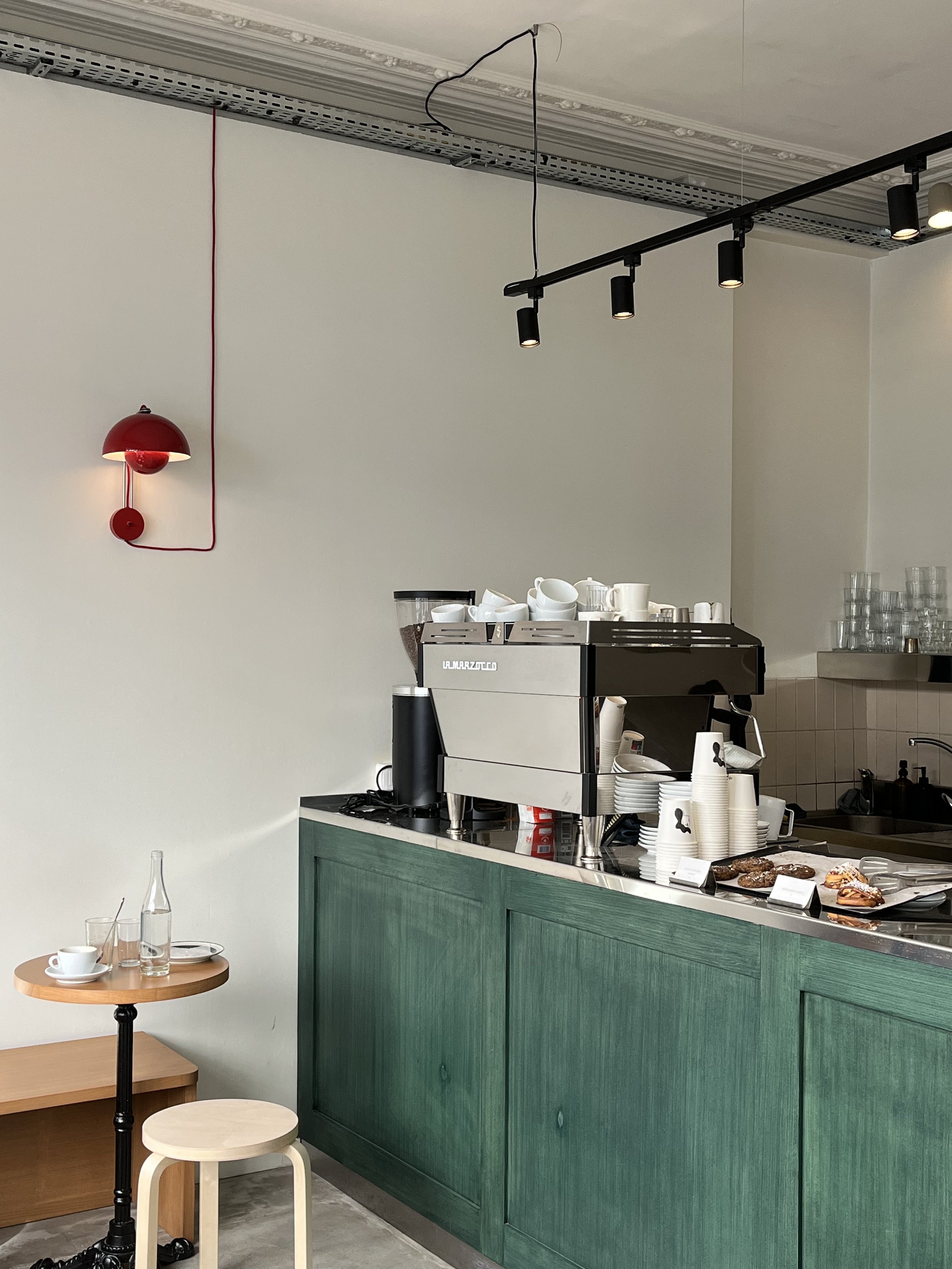 Interior of a coffee shop or café, showing a green counter with a coffee machine, cups, and pastries, along with some seating including a small wooden table with a cafe tray, cups, and water bottles.