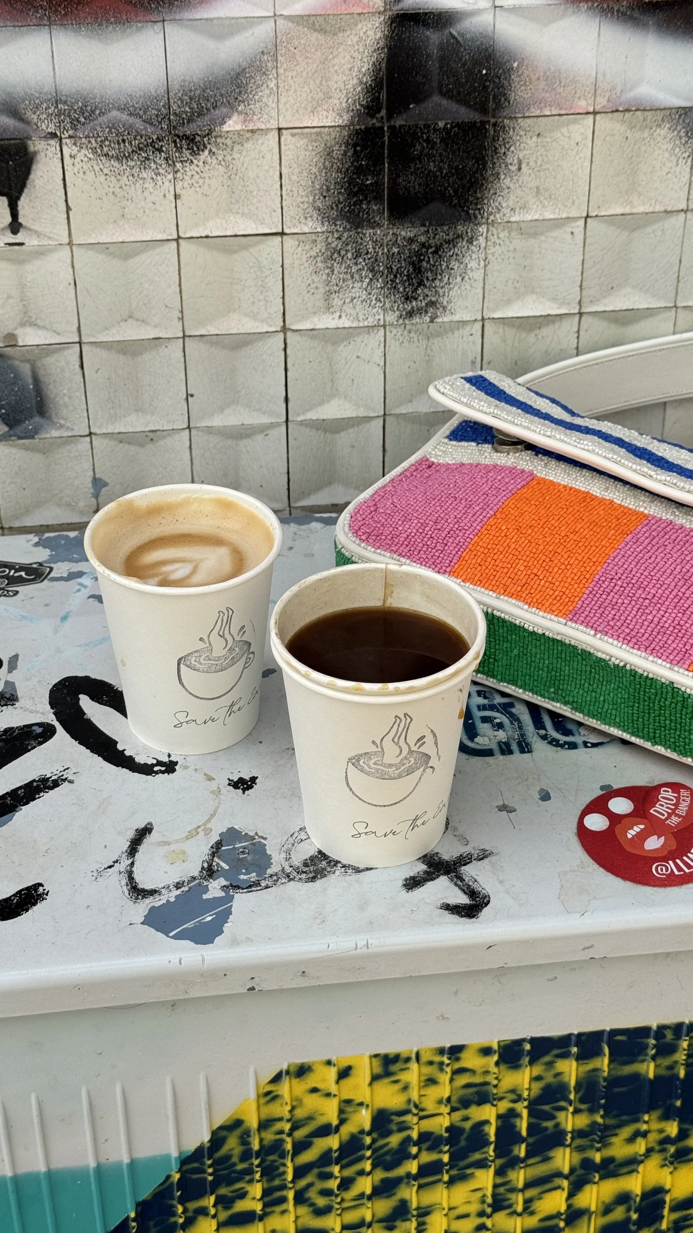 Two paper cups with hot beverages, one appears to be coffee and the other hot chocolate, on a worn, graffiti-covered table with a colorful beaded wallet and a tiled wall background.