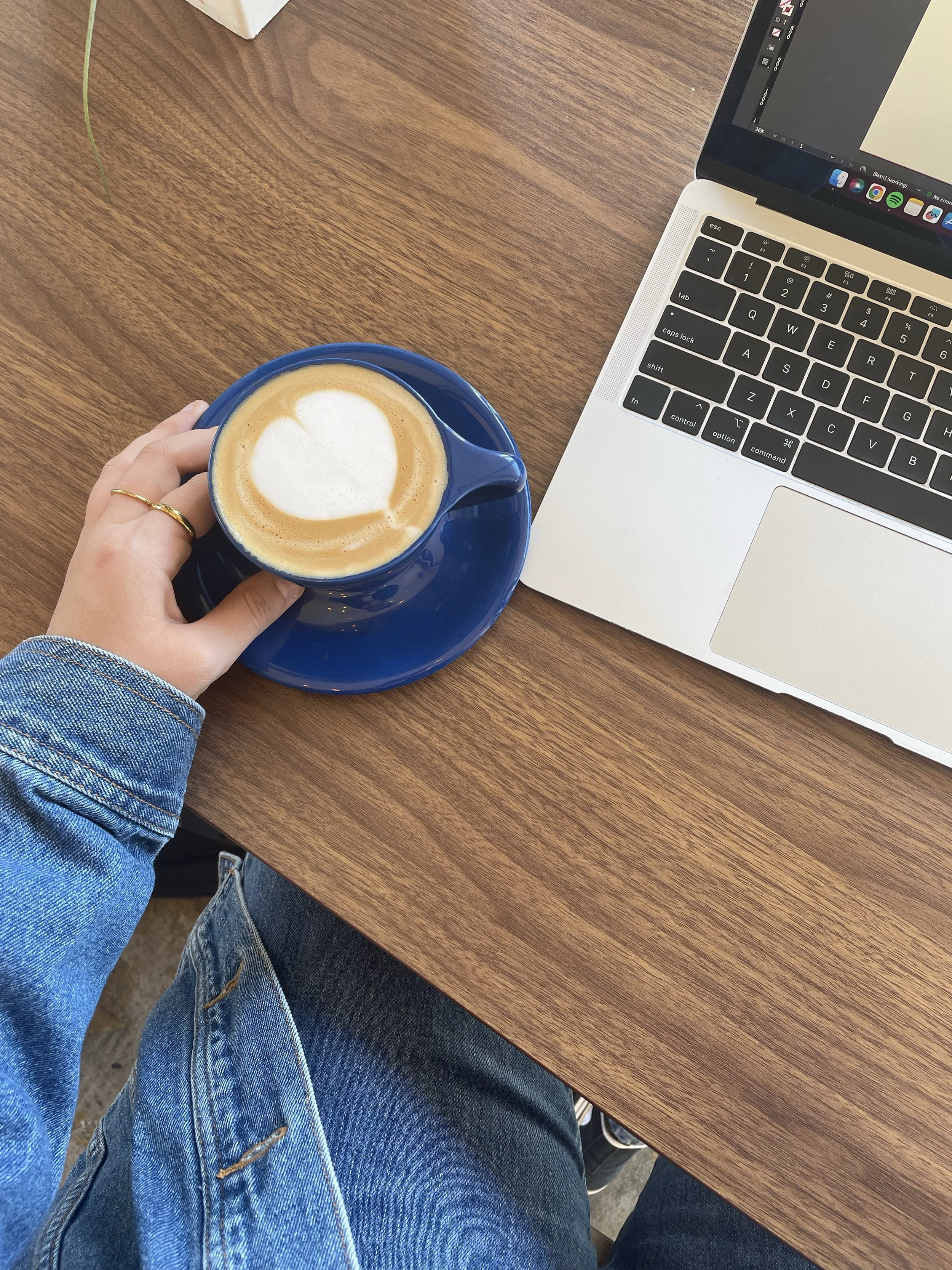 Person wearing jeans sitting at a wooden table, holding a blue cup of coffee with foam art, with a laptop open on the table.