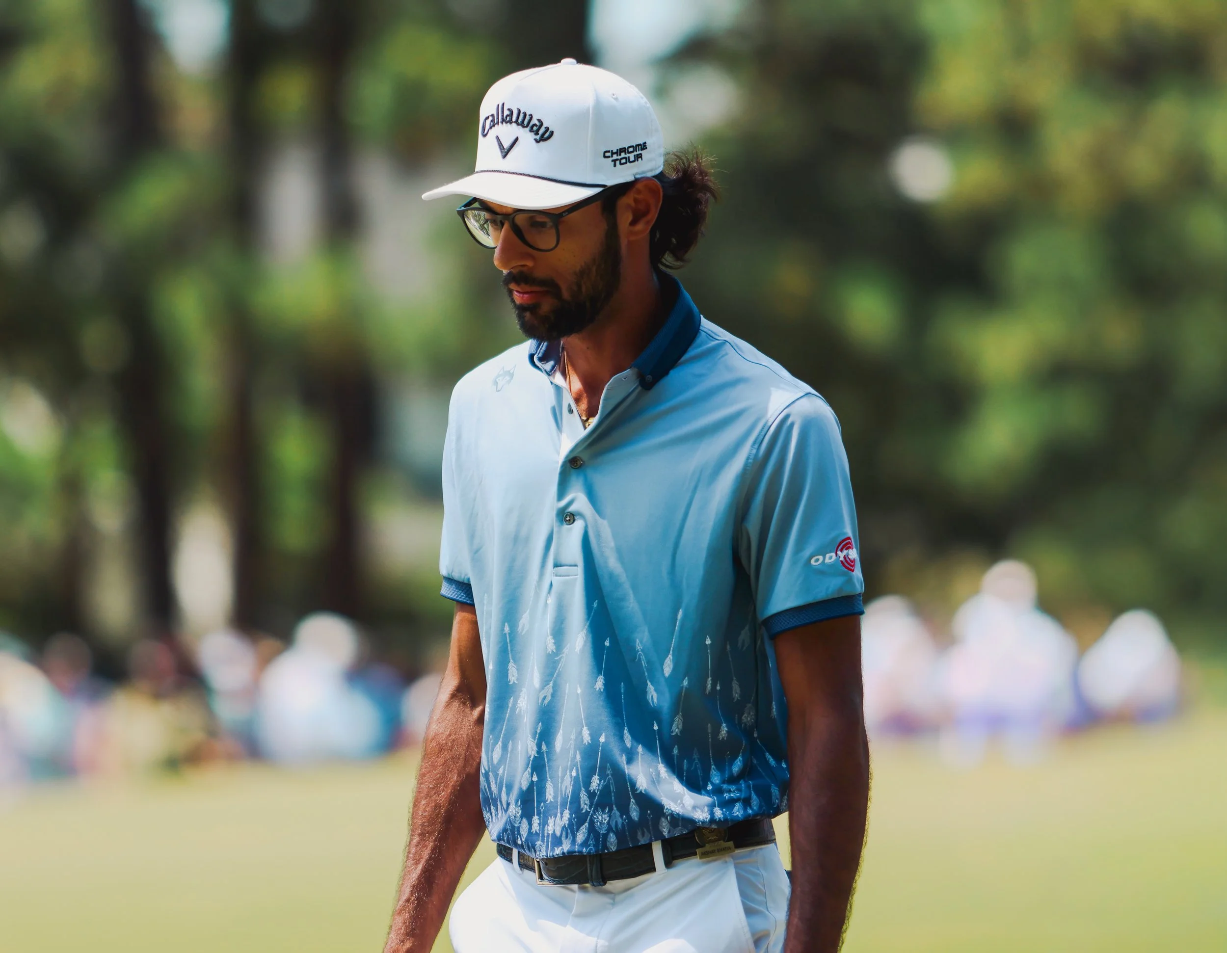 A man wearing glasses and a Callaway golf cap stands outdoors on a golf course, dressed in a light blue collared shirt with fish patterns and white golf shorts, with a blurred background of trees and spectators.