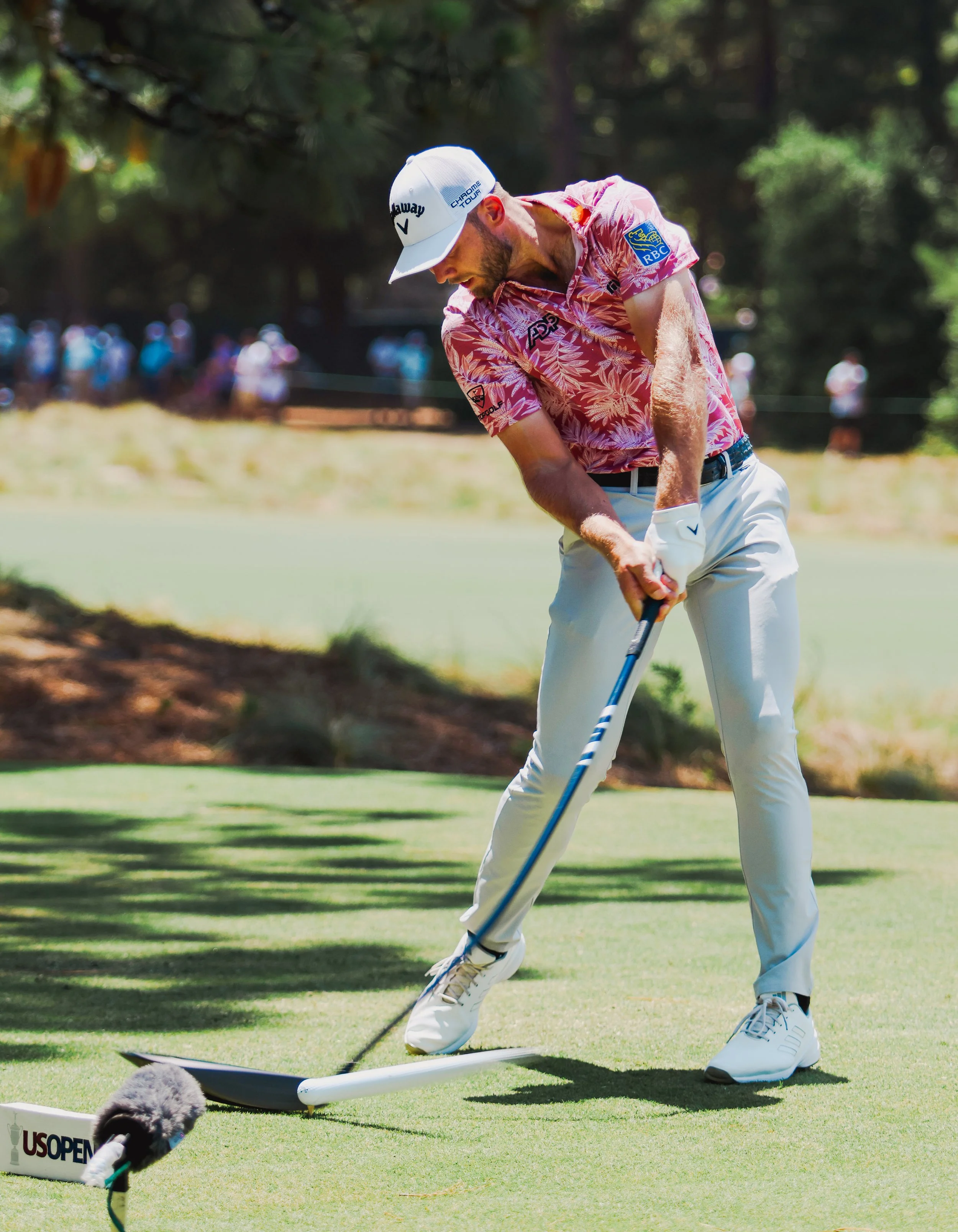 A golfer on the green surface, preparing to putt, wearing a pink Hawaiian shirt, white pants, and a white cap. The scene is outdoors with trees and spectators in the background during a sunny day.
