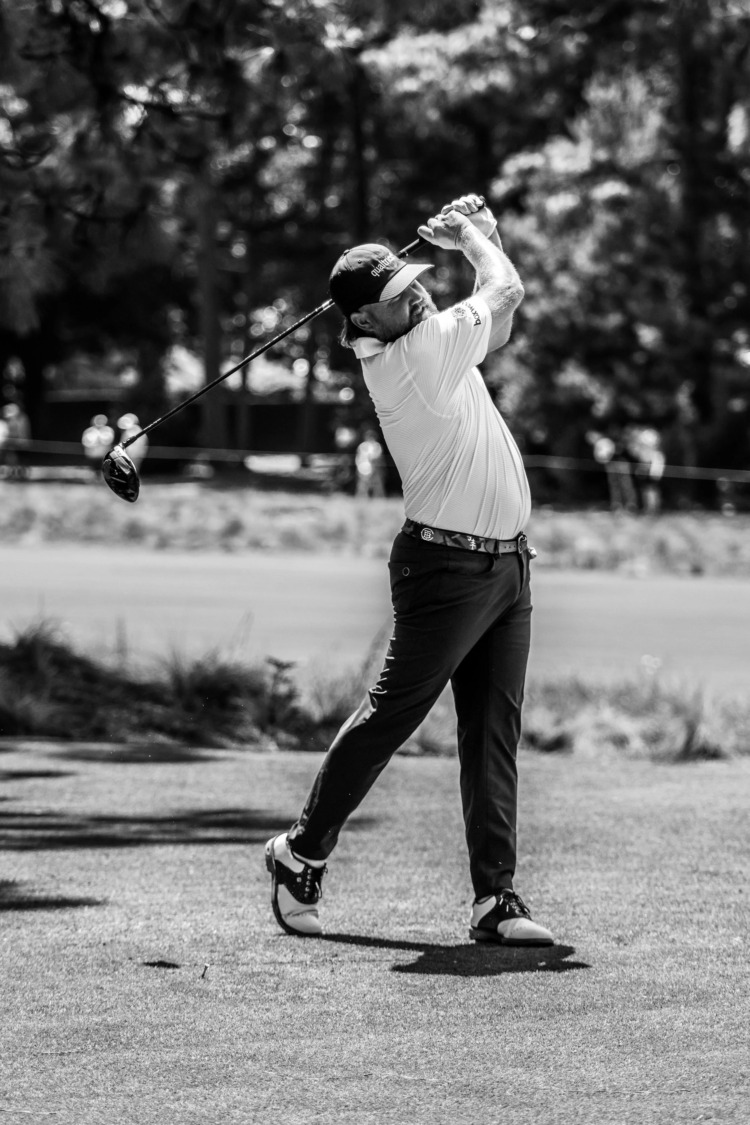 A male golfer wearing a cap and polo shirt swinging a golf club on the course, with trees and a few people visible in the background.