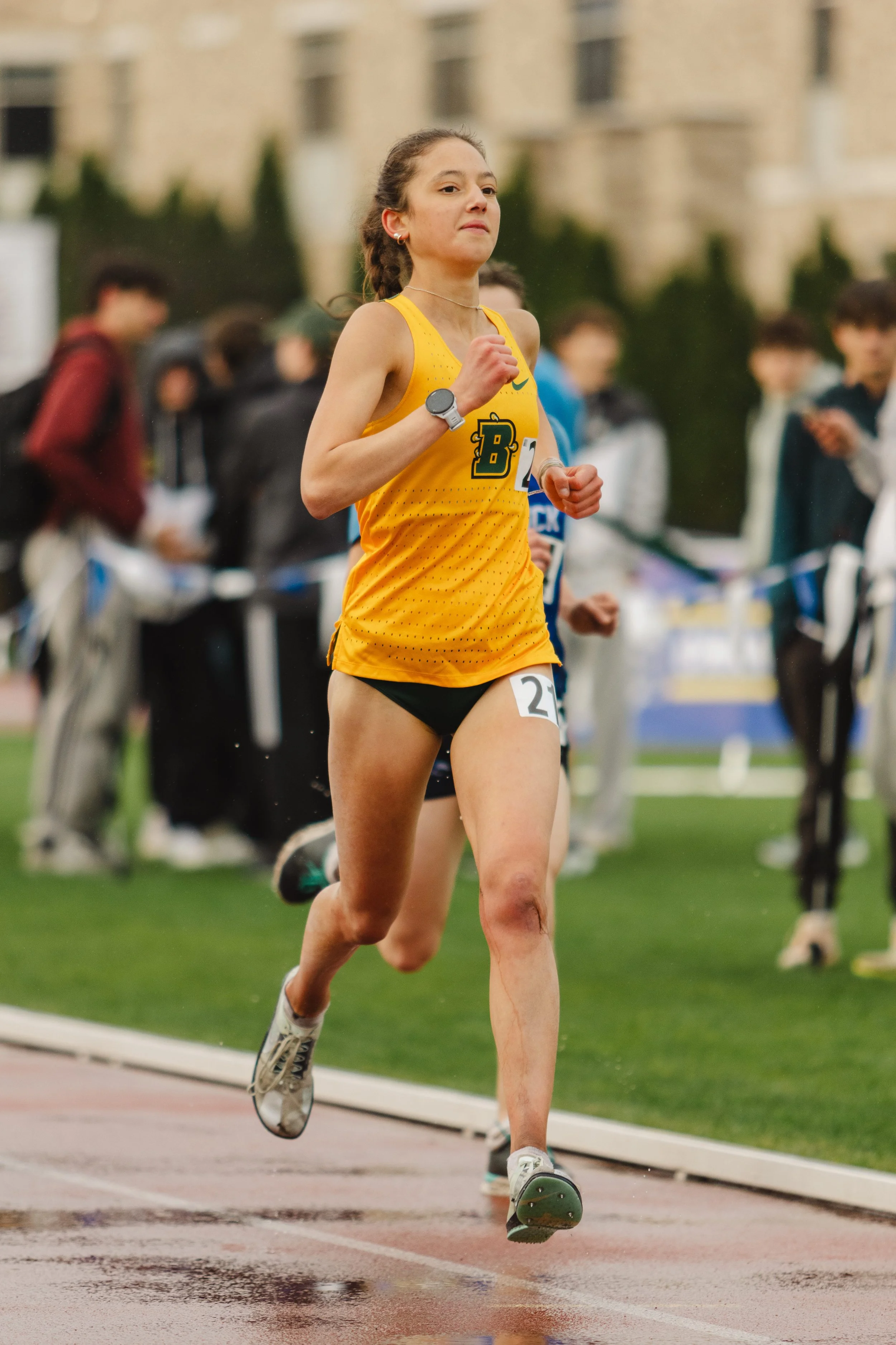 A female runner in a yellow sports top and black shorts running on a track with a determined expression. She is wearing a fitness watch, earrings, and a necklace. There are spectators and other runners in the background, along with a building and som