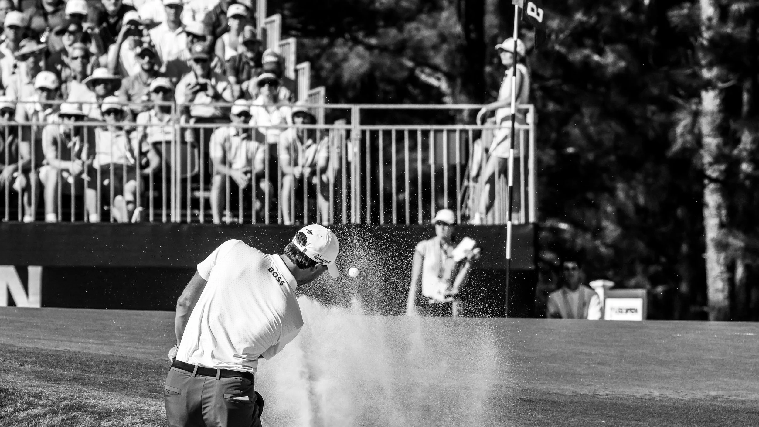 A golfer in a white shirt and cap is taking a shot from a sand trap on a golf course, with spectators watching behind a fence in the background.