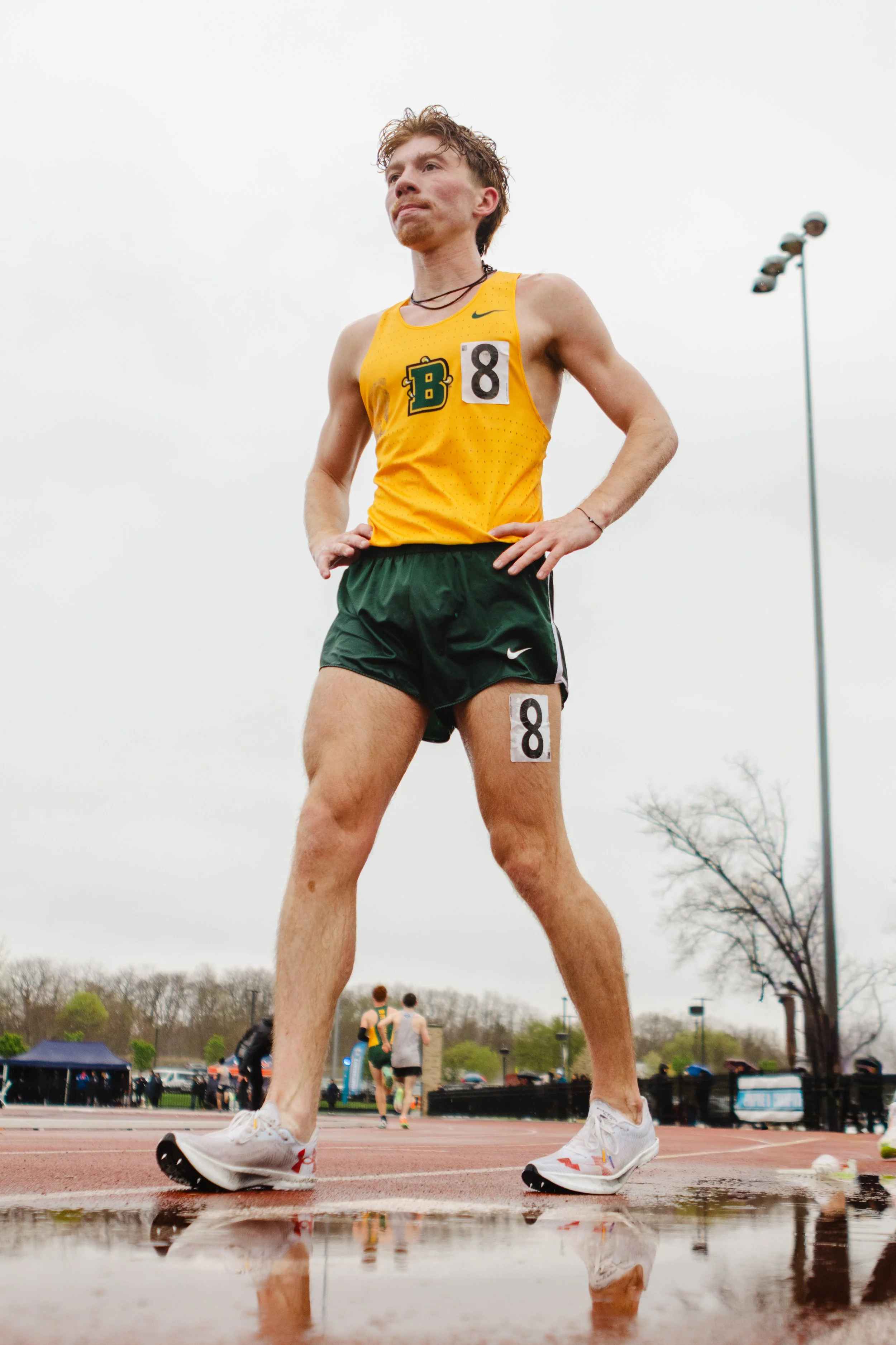 A male athlete wearing a yellow and green running uniform with bib number 8, standing on a wet track with water puddles, looking forward after a race. He is wearing white running shoes and has a determined expression. The background shows other runne