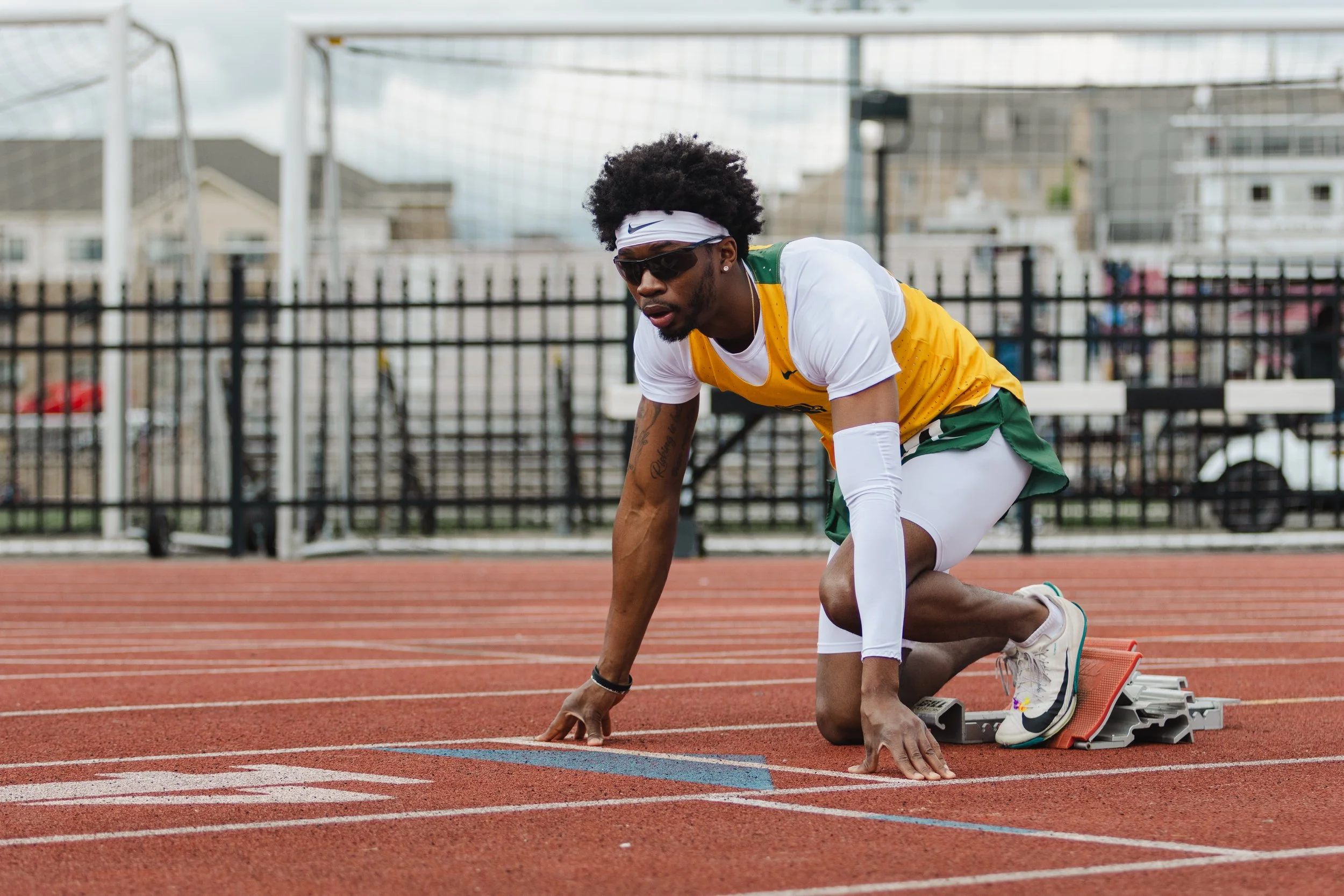 A male sprinter wearing sunglasses, a white headband, and a yellow and green athletic uniform crouches at the starting block on a red track, preparing for a race with a residential neighborhood in the background.