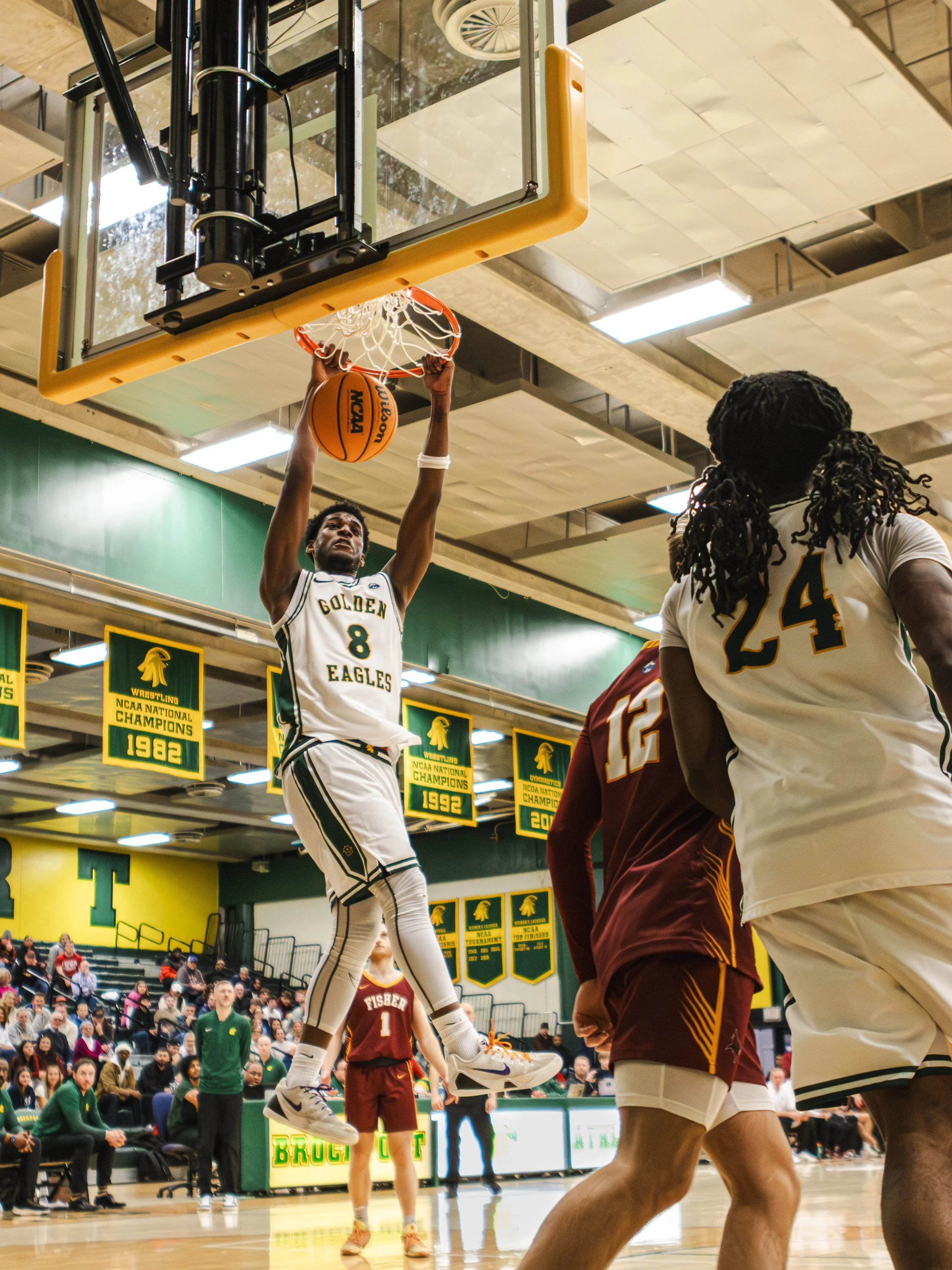 A basketball player in a white and green uniform from SUNY Brockport jumps to dunk the ball during a game in a gym with banners hanging from the ceiling and spectators in the background.