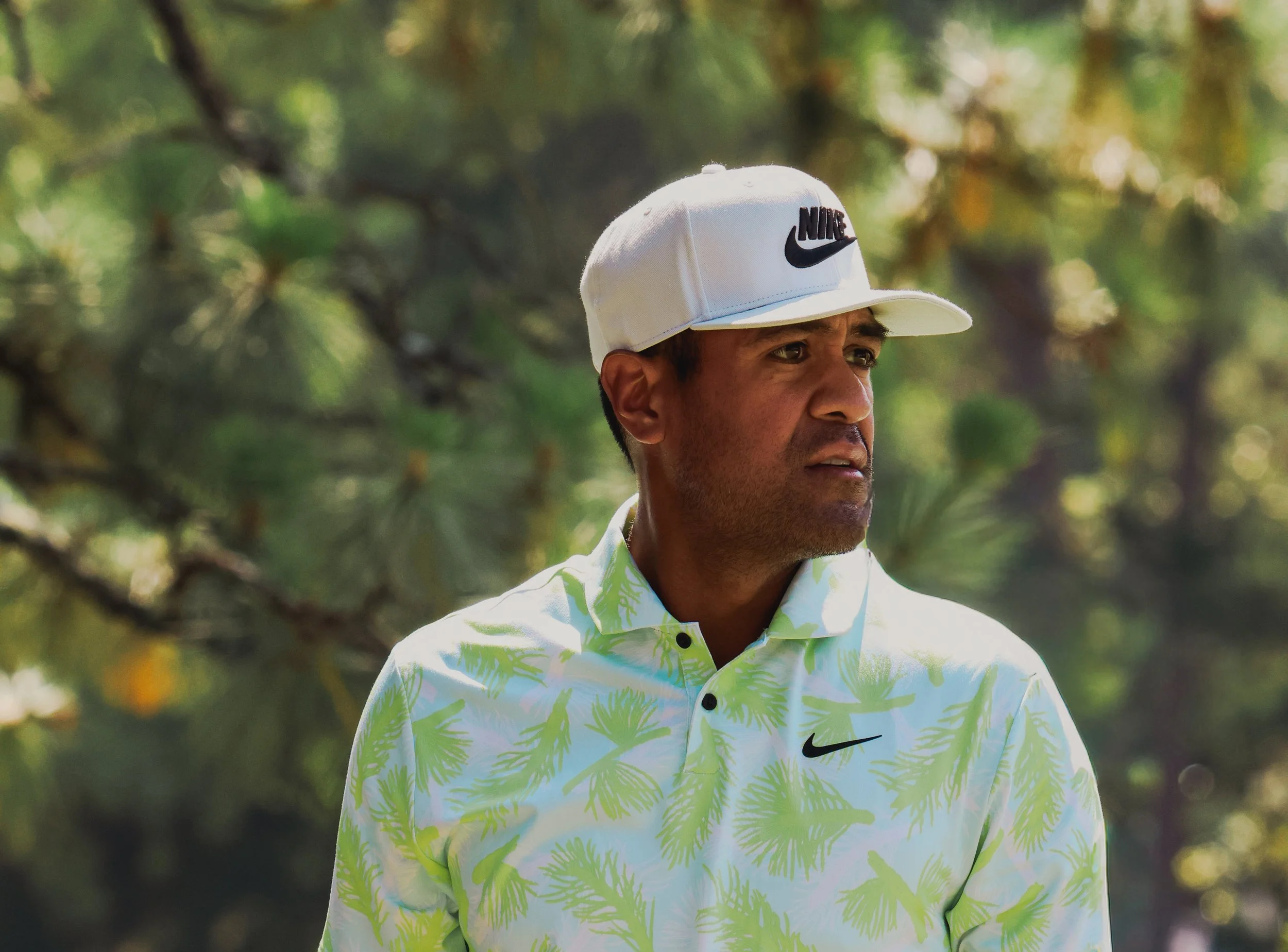 A man wearing a white Nike cap with the logo and a white shirt with green palm leaf patterns stands outdoors with blurred trees in the background.