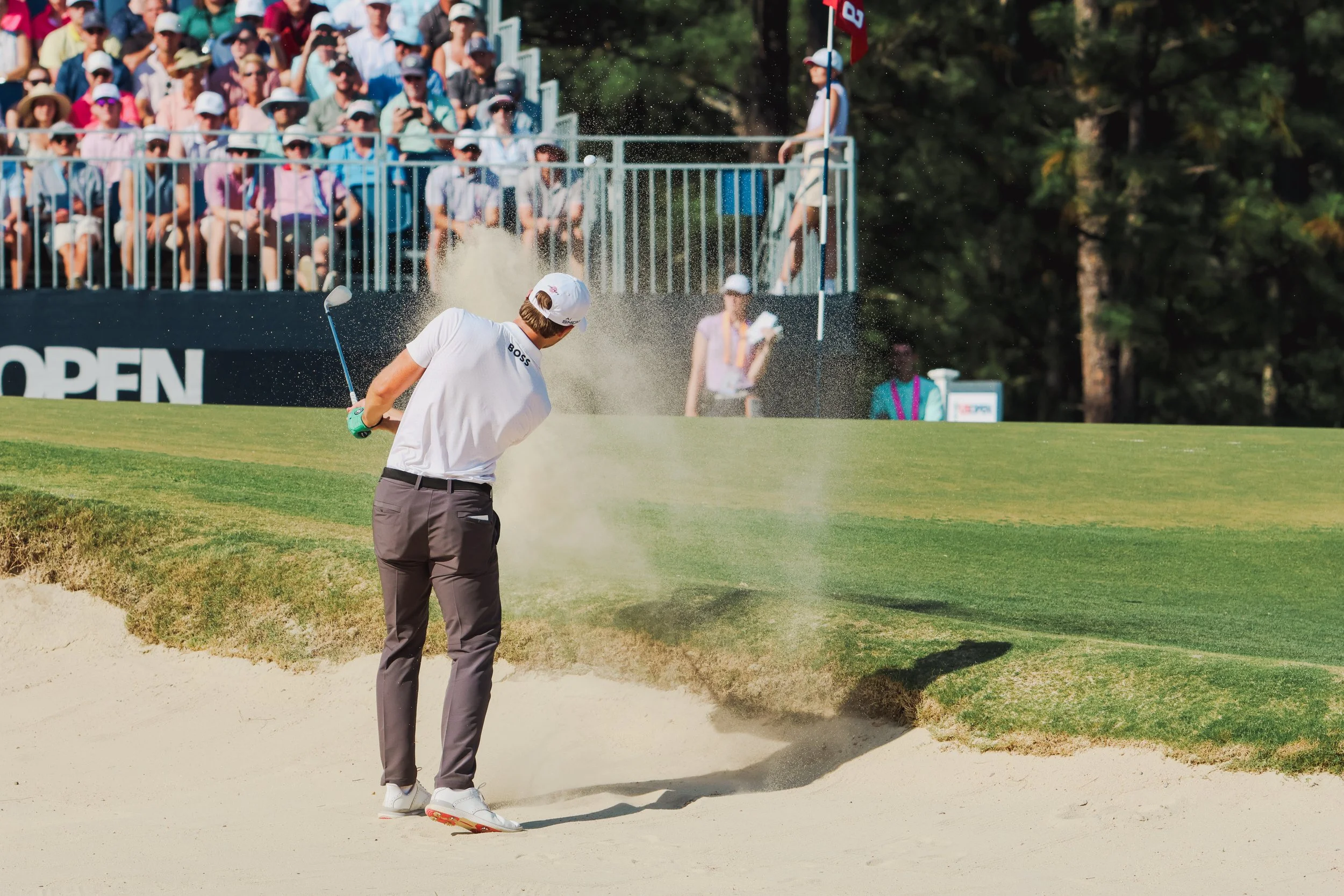 A golfer standing in a sand trap bunker on a golf course, with spectators seated behind a fence watching the game.