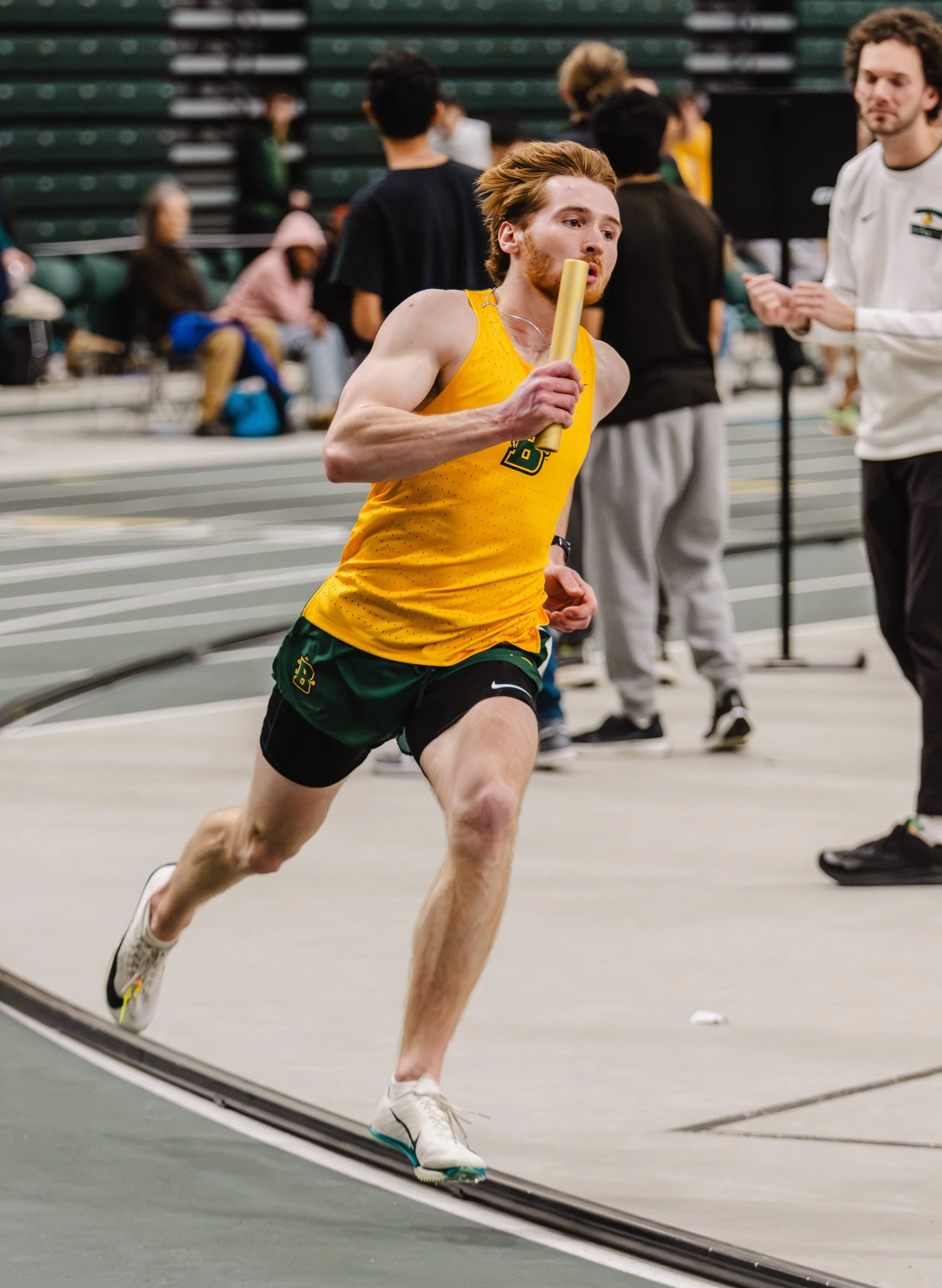 A male athlete running indoors during a track event, holding a relay baton, wearing a yellow and green uniform from SUNY Brockprot, with spectators and other participants in the background.