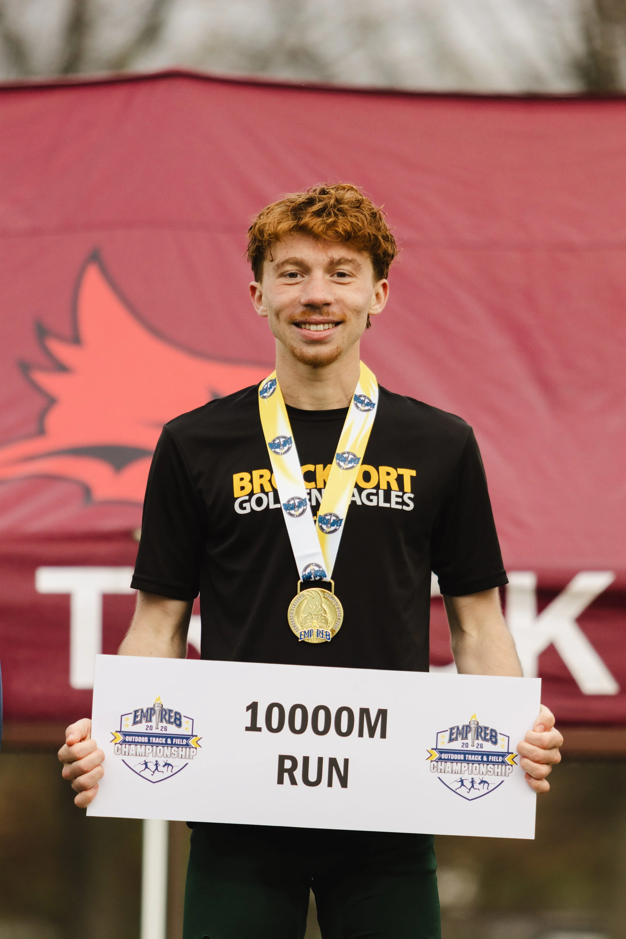 A young man with curly hair and a medal around his neck, standing outdoors, holding a large sign that reads '10000M RUN' at the Empire Outdoor Track & Field Championship.
