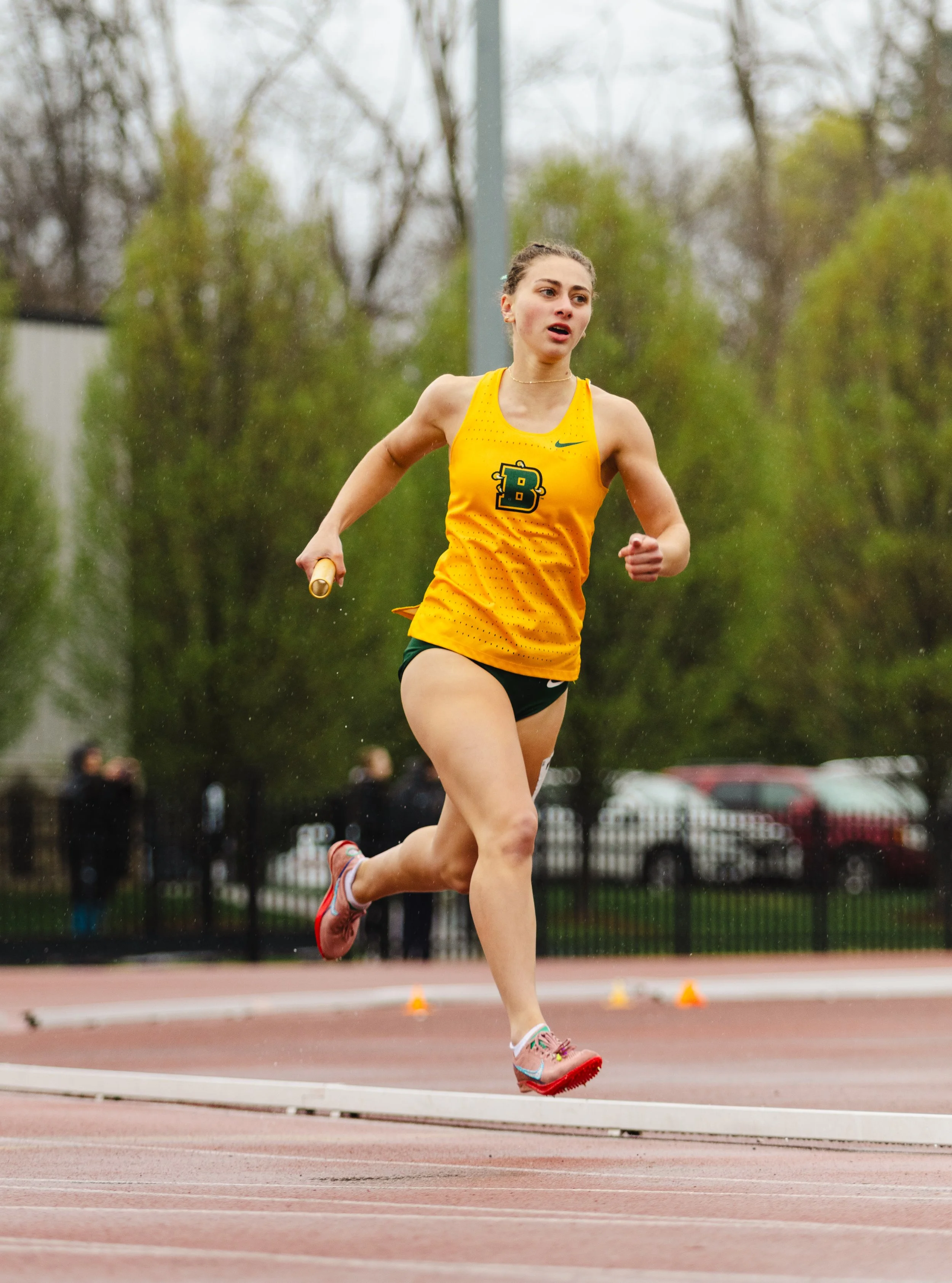 A young female runner in a yellow running uniform with green accents, running on a track during a race, holding a relay baton, with trees and spectators in the background.